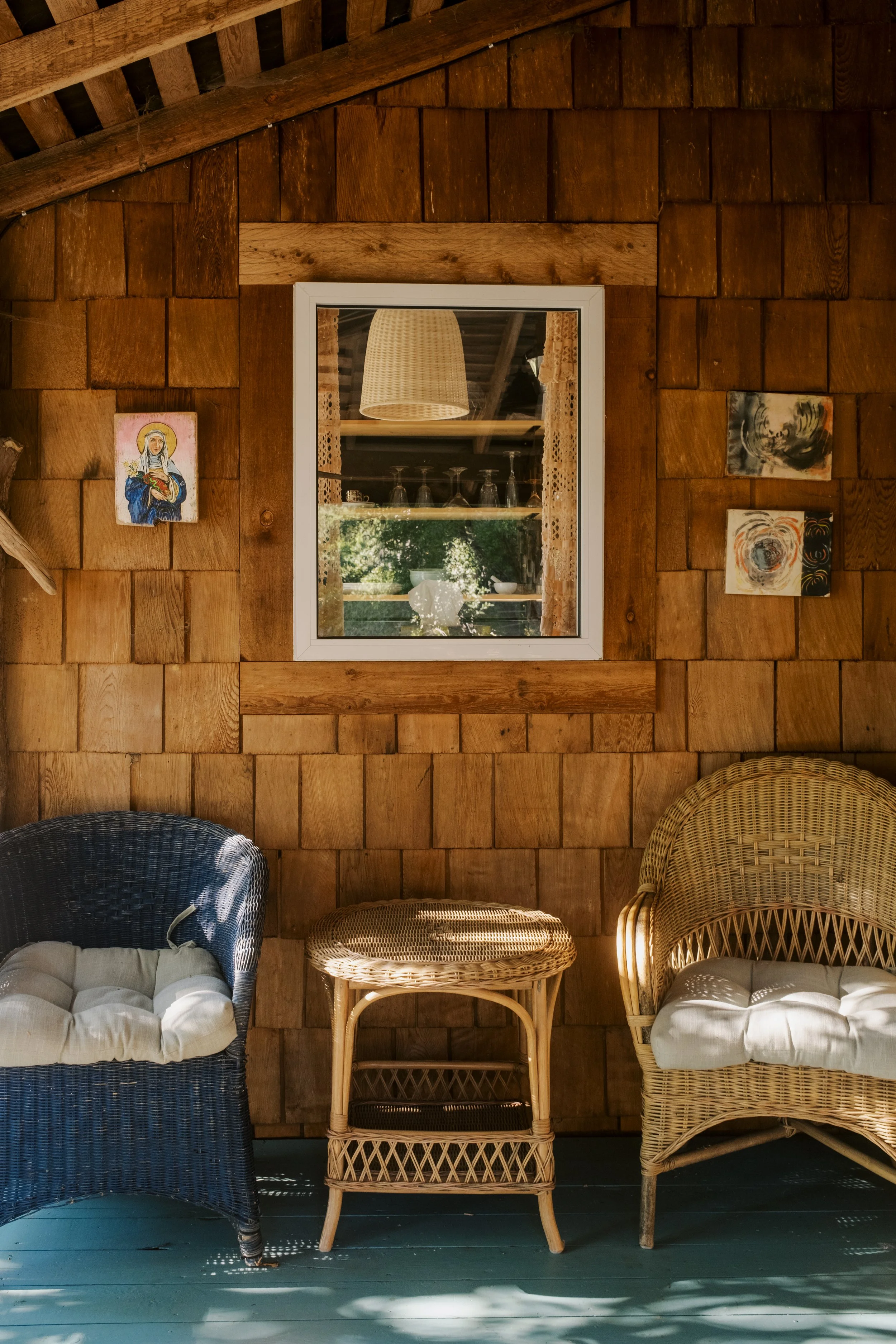 A cozy wooden porch with wicker and rattan chairs, a small side table, and a window with lace curtains. Artwork hangs on the wood-paneled wall.