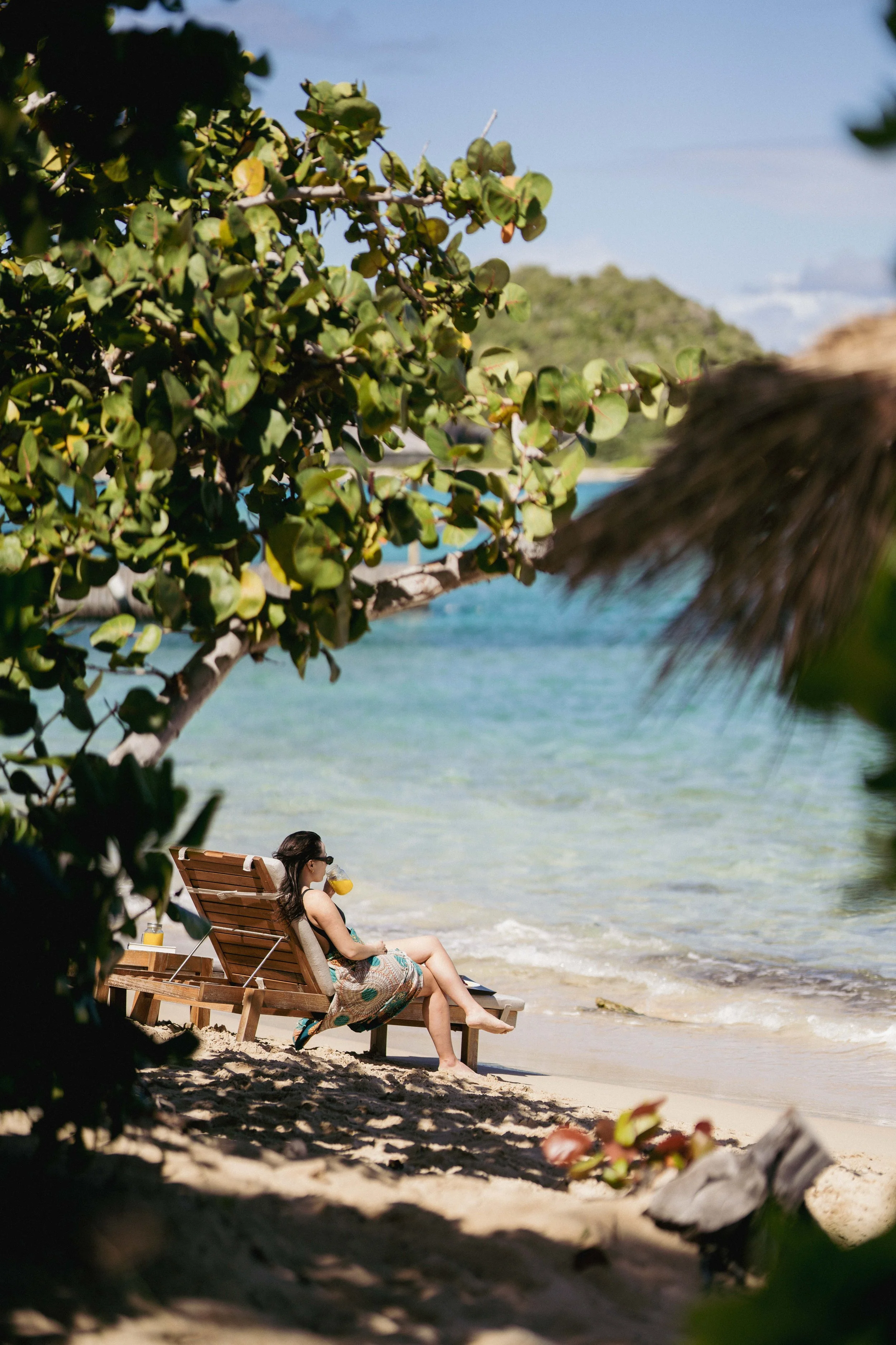 A woman relaxing on a wooden lounge chair on a sandy beach, sipping a drink through a straw, with lush green foliage and clear blue water in the background.