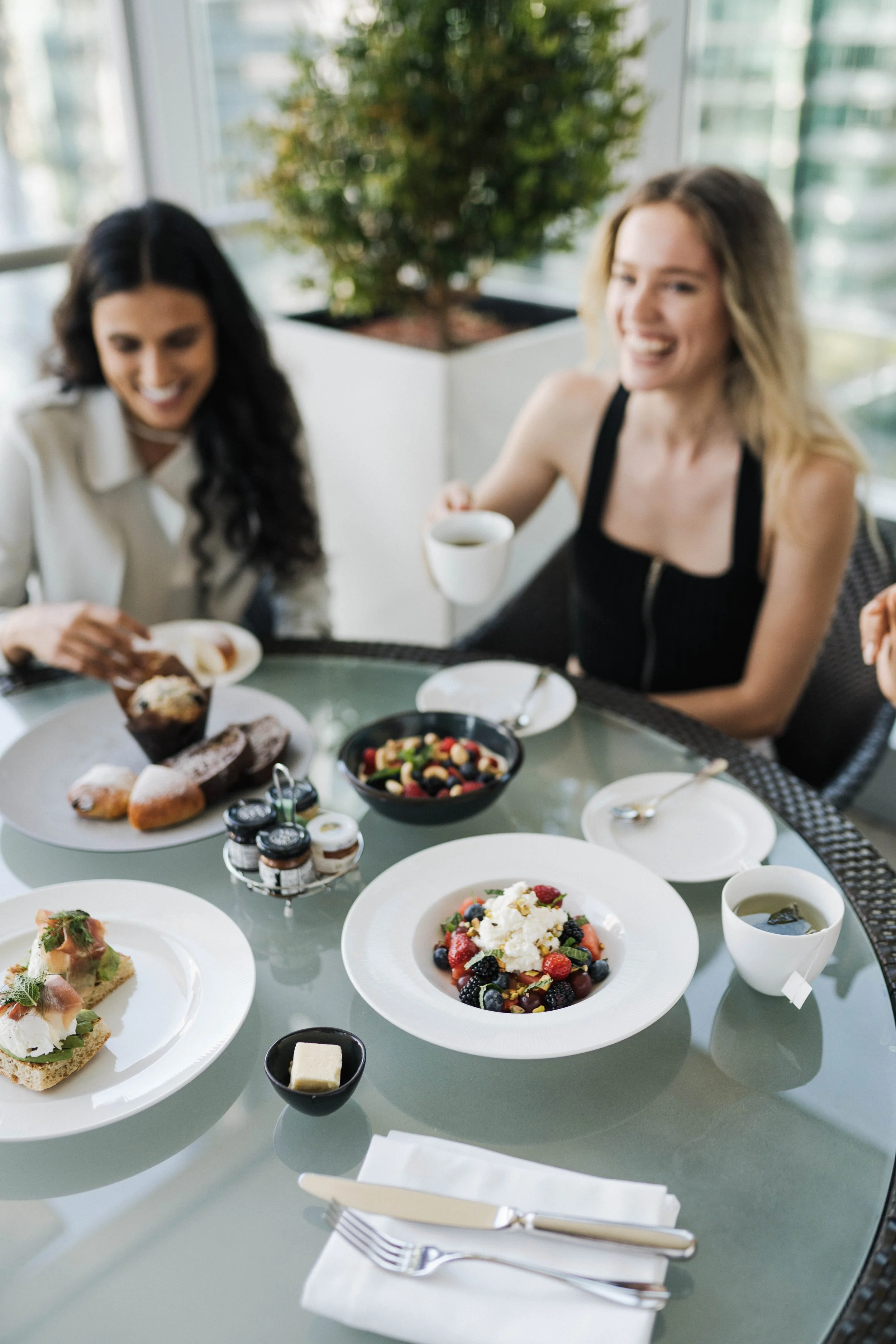 Two women enjoying a meal with various desserts and drinks at a glass table in a bright, modern setting.