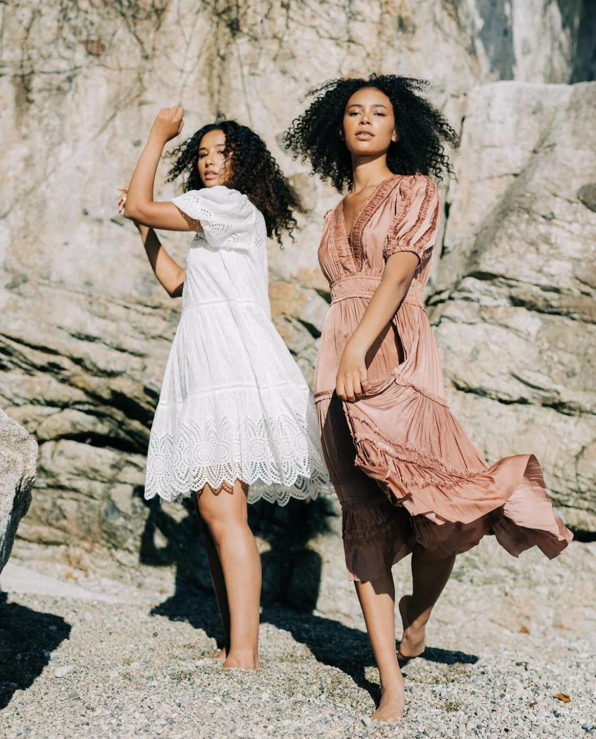 Two women with curly hair posing on rocks at the beach, wearing flowy dresses, one white and the other pink.