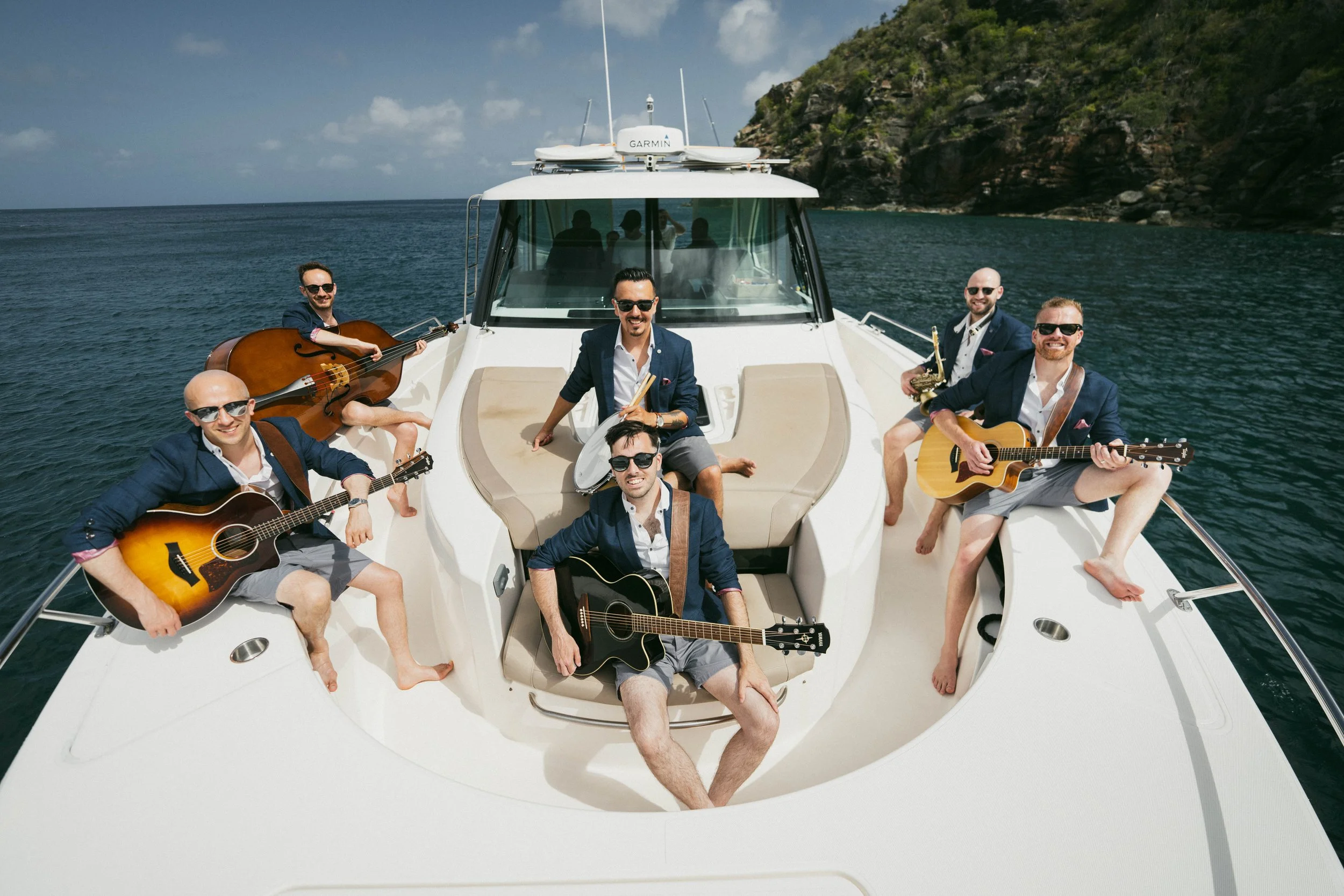 Group of six men on a yacht, playing guitars, drums, and a saxophone, with a scenic ocean and rocky hillside in the background.