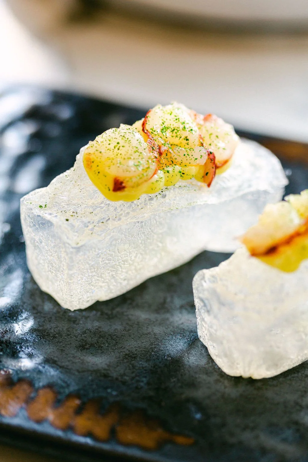 Close-up of two pieces of ice topped with a small portion of creamy yellow and orange seafood salad garnished with finely chopped herbs on a dark surface.