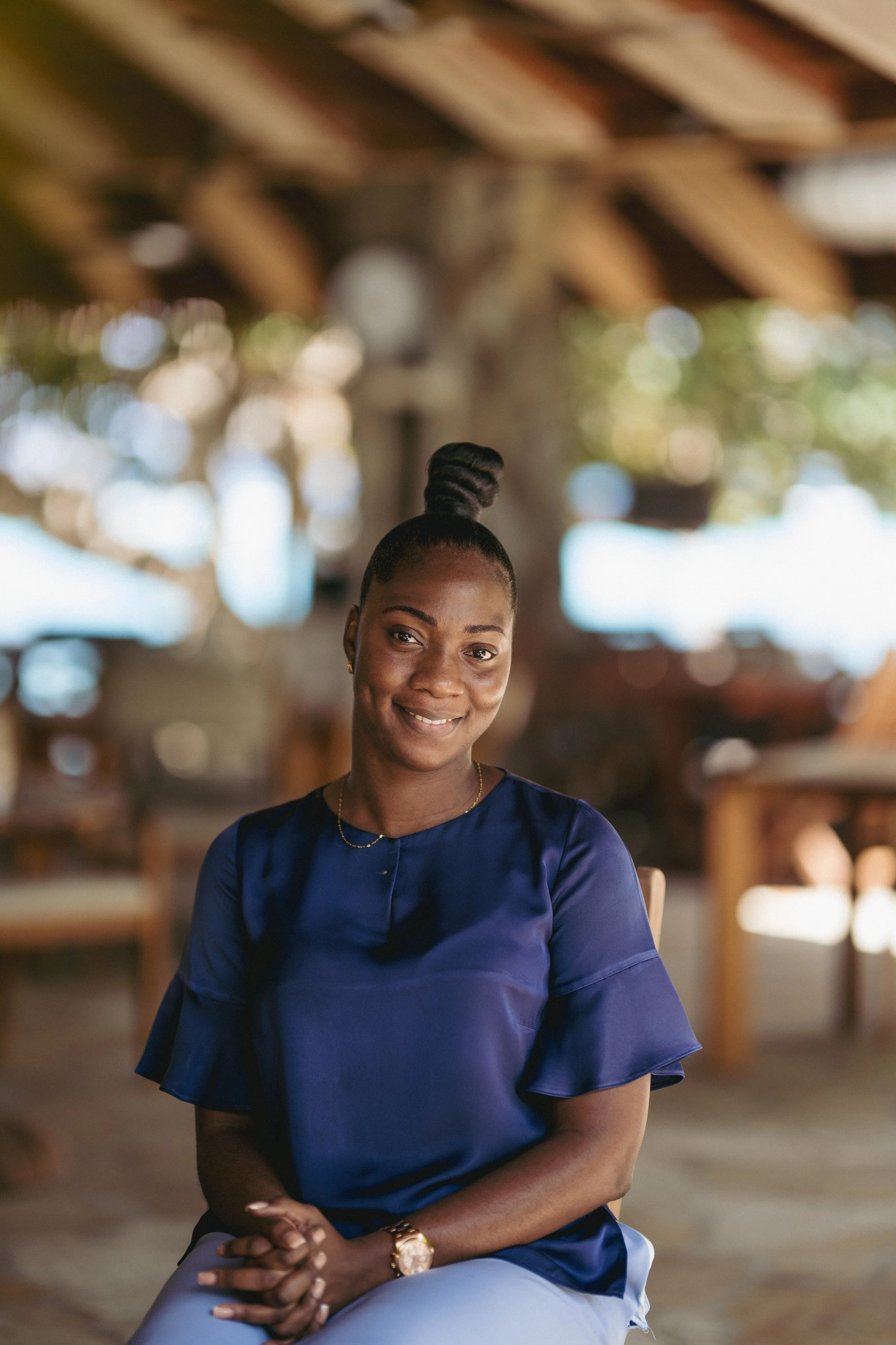 A woman sitting outdoors in a shaded area, wearing a navy blue blouse with ruffled sleeves, with her hands clasped on her lap, smiling at the camera.