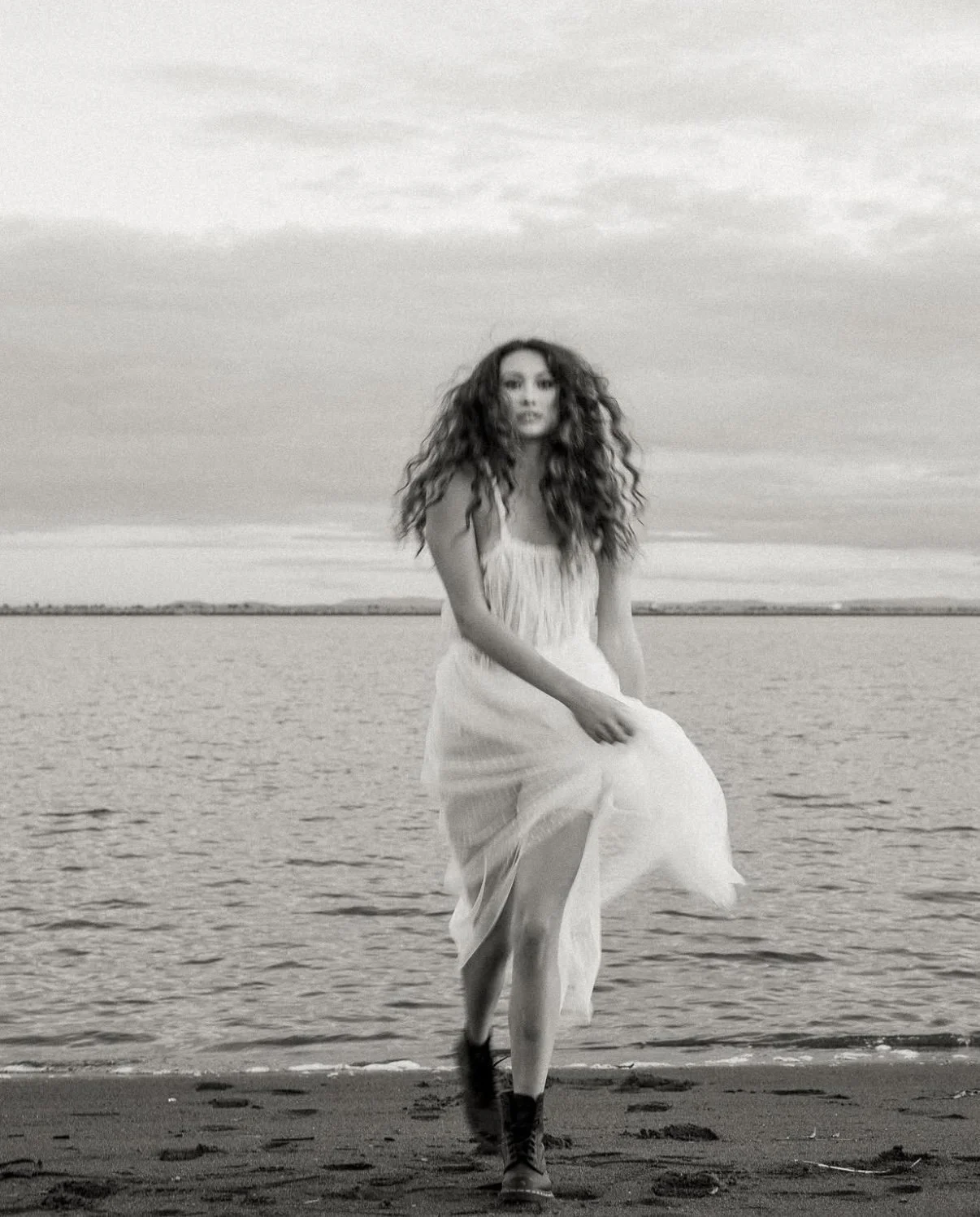 A woman with long, curly hair in a light dress and boots walking on a beach.