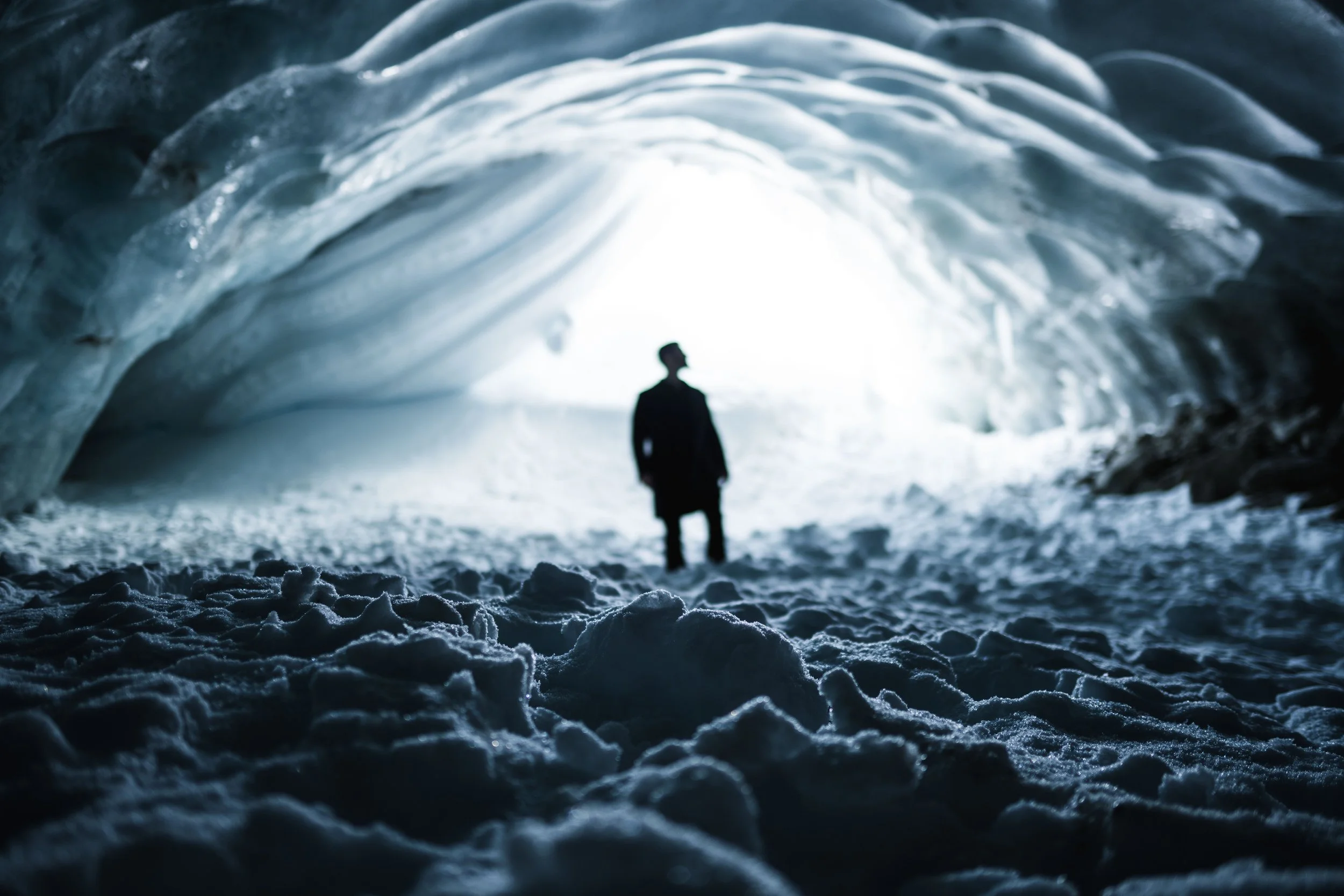 A person standing inside an icy ice cave with snow on the ground, silhouetted against a bright opening at the end of the cave.