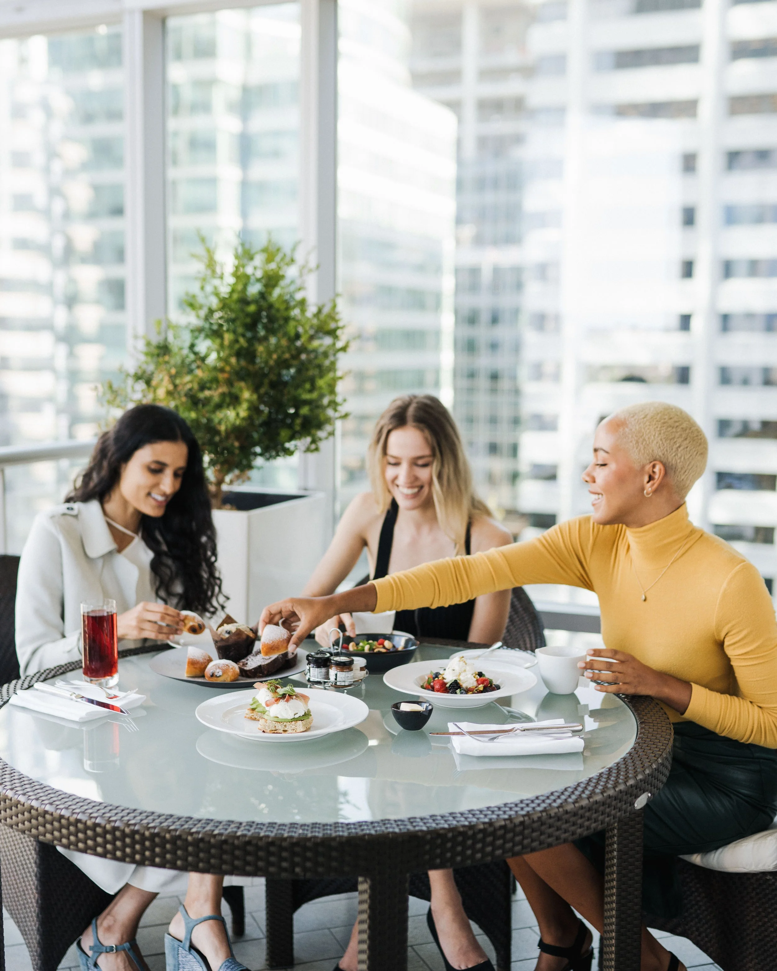 Three women gathered around a table with desserts and drinks in a bright indoor space with large windows and city views.