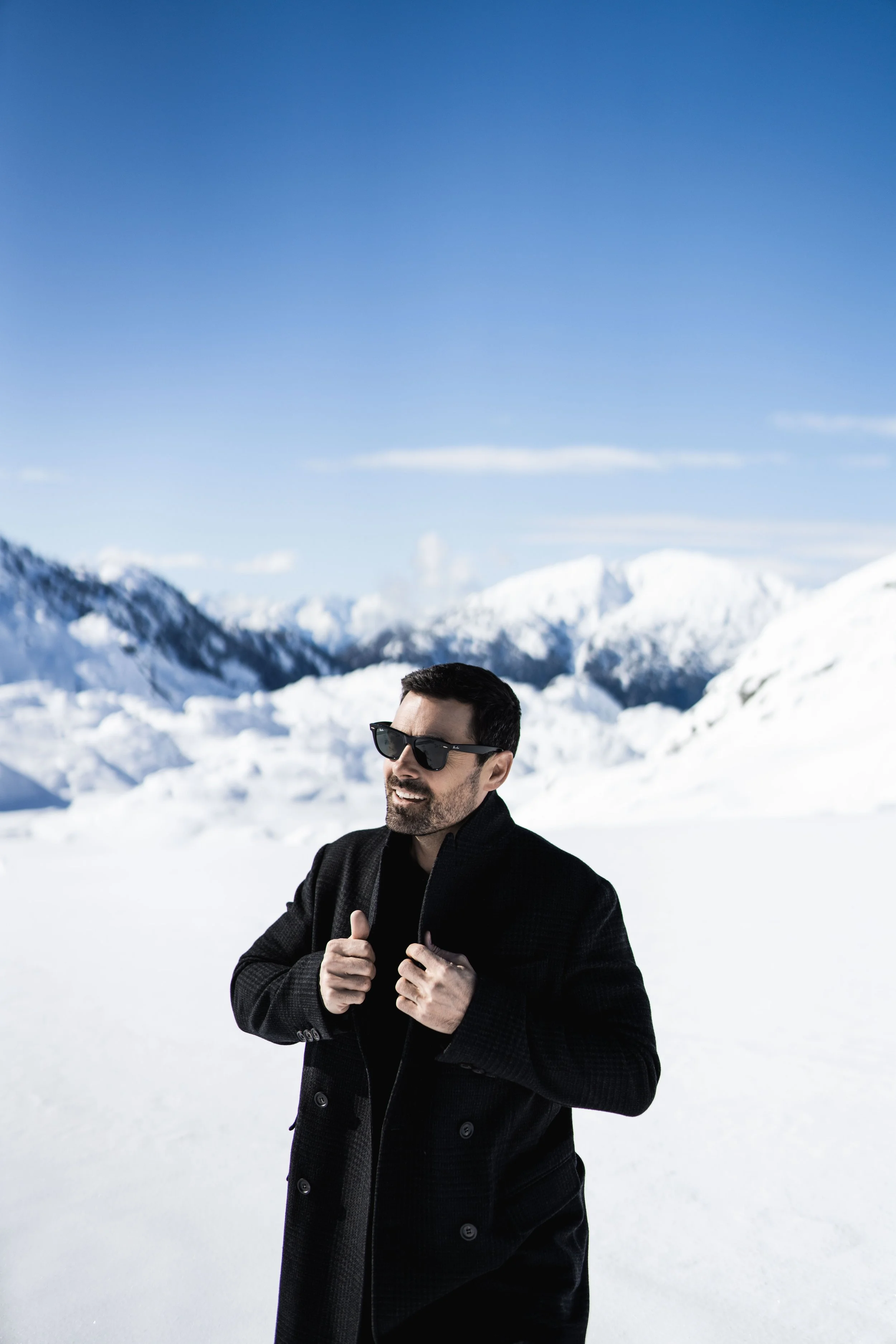 A man in black winter coat and sunglasses standing in snowy mountain landscape with blue sky and snow-covered mountains in the background.
