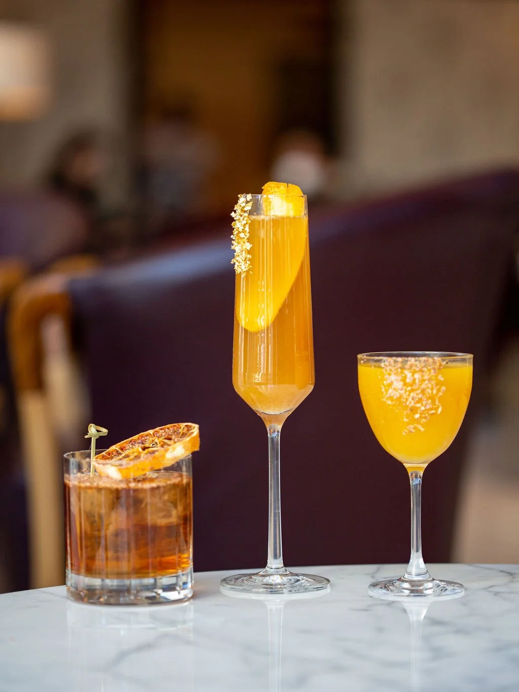 Three cocktails on a white marble table with a blurred background. The left drink is amber-colored with a dried orange slice garnish, the middle drink is a tall yellow cocktail in a flute glass with a lemon wedge and white sugar rim, and the right dr