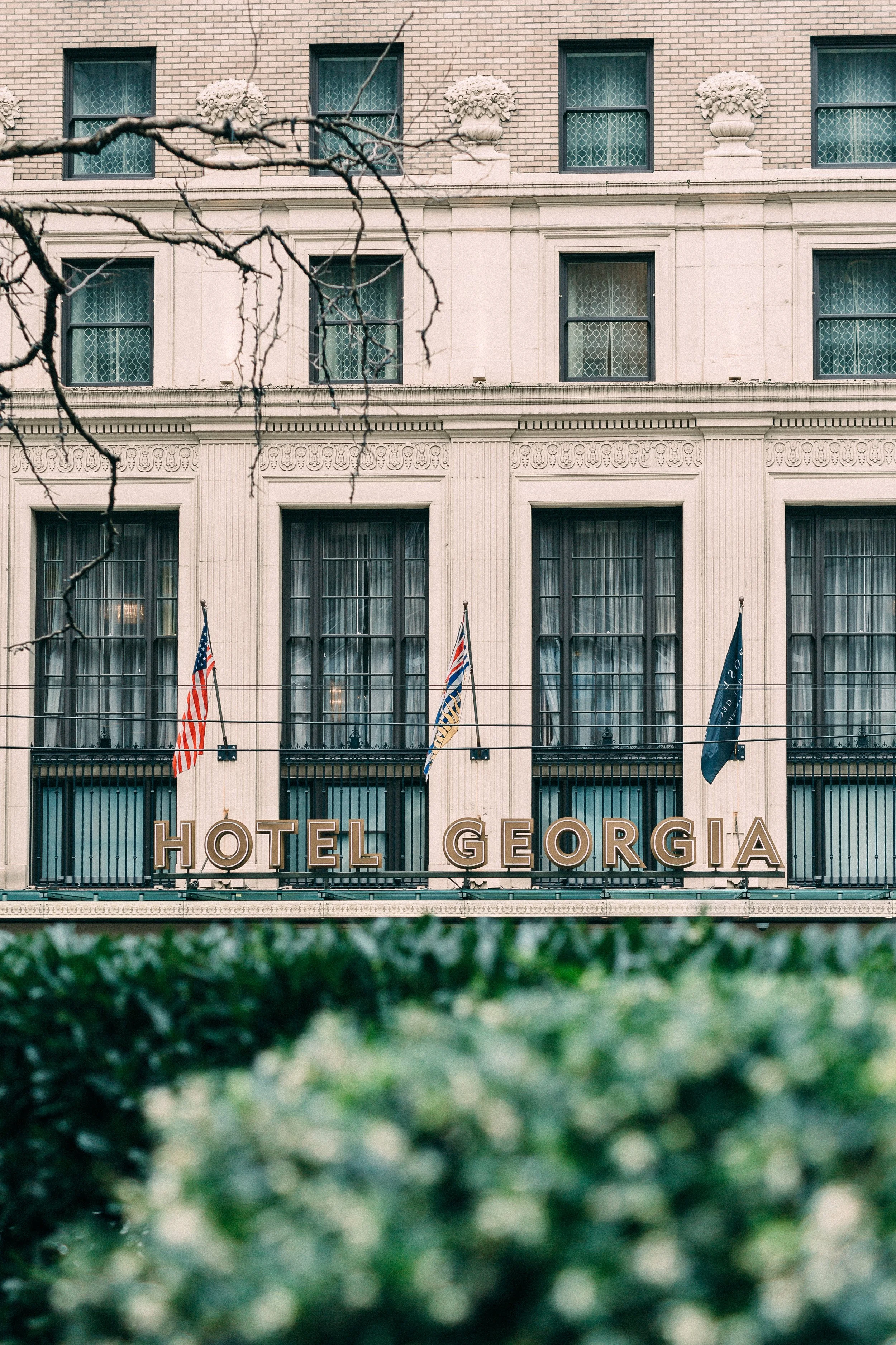 The exterior of the Hotel Georgia with its sign and flags, framed by some tree branches and greenery in the foreground.