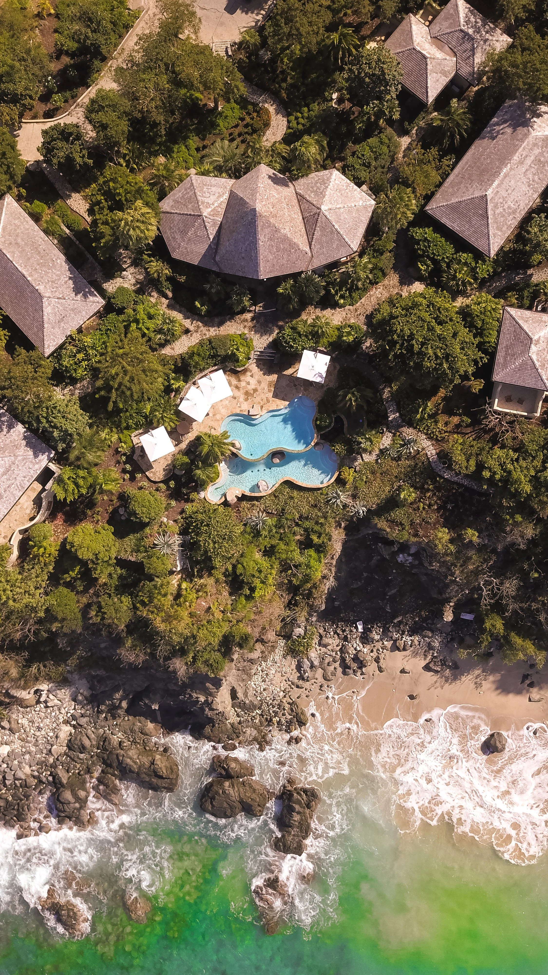 An aerial view of a tropical resort with a swimming pool, surrounded by lush greenery, and close to the beach with rocks and ocean waves.