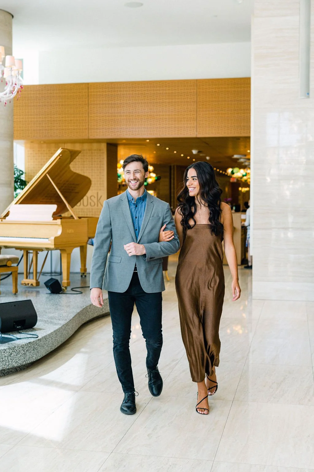 A man and woman walking arm in arm in an elegant hotel lobby, smiling, with a grand piano in the background and festive decorations.