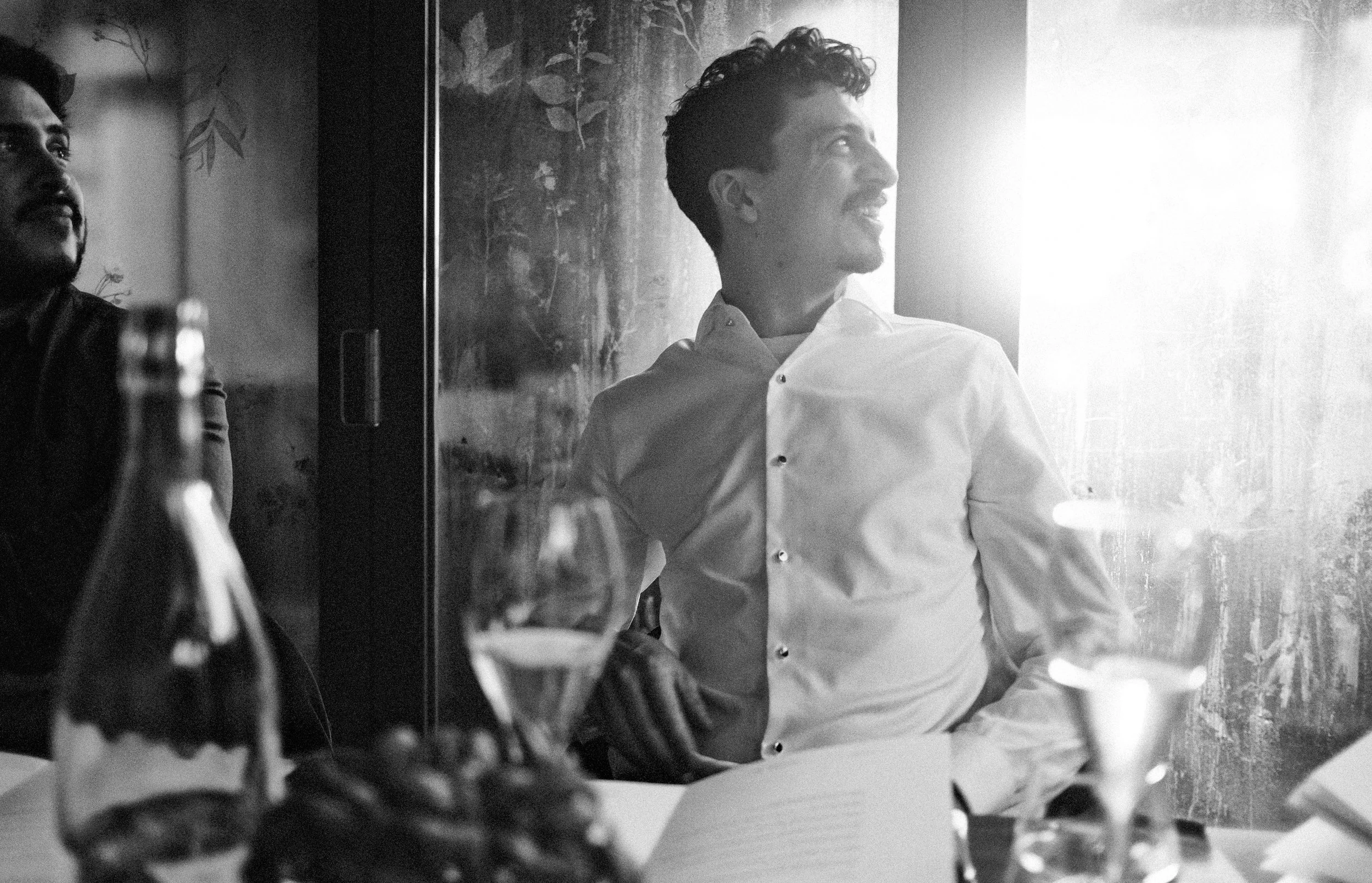 A young man in a white dress shirt smiling while sitting at a table in a dimly lit restaurant, with sunlight streaming from the right side of the image.