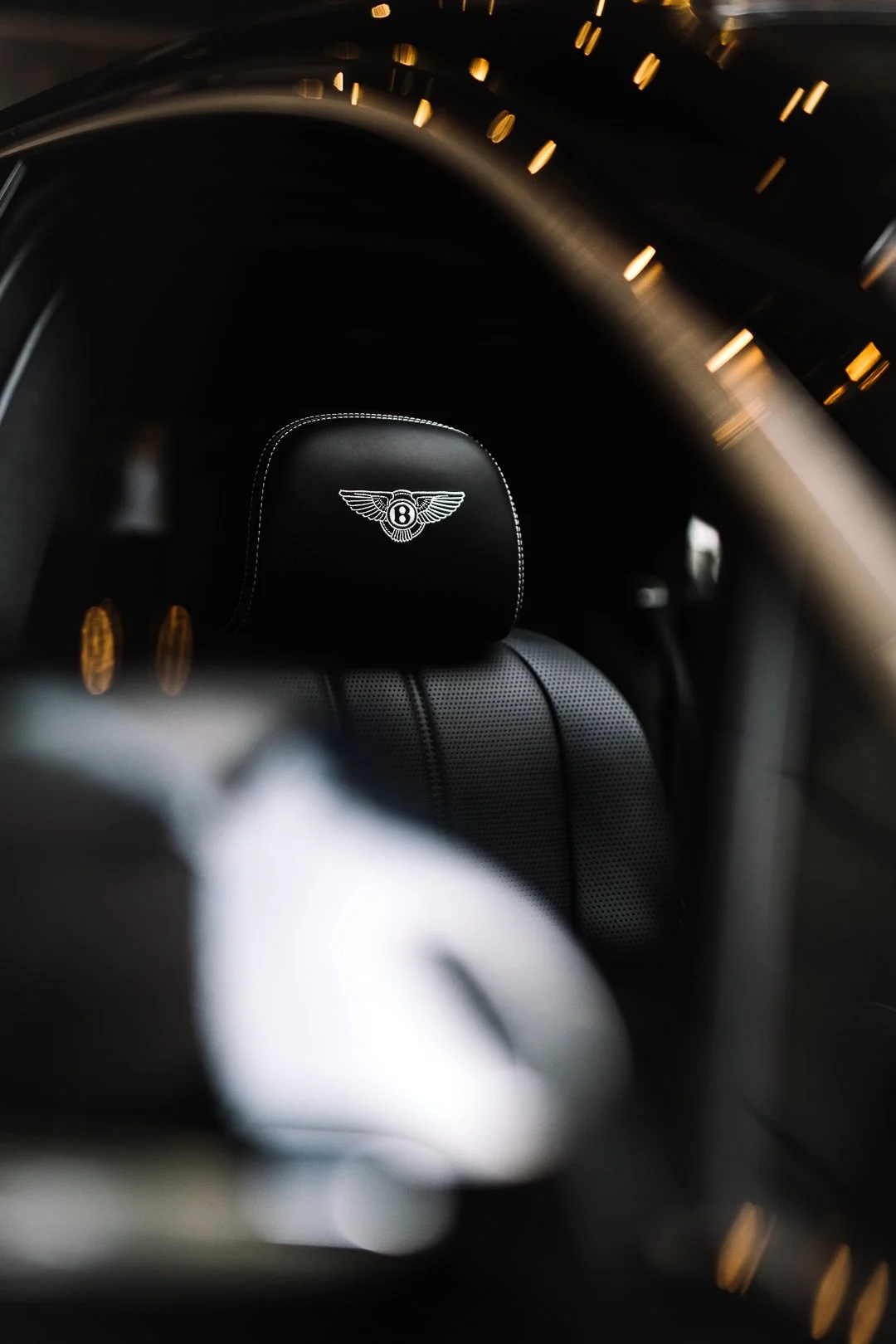 Close-up of a Bentley car seat headrest with a Bentley emblem, surrounded by blurred interior and lighting reflections.