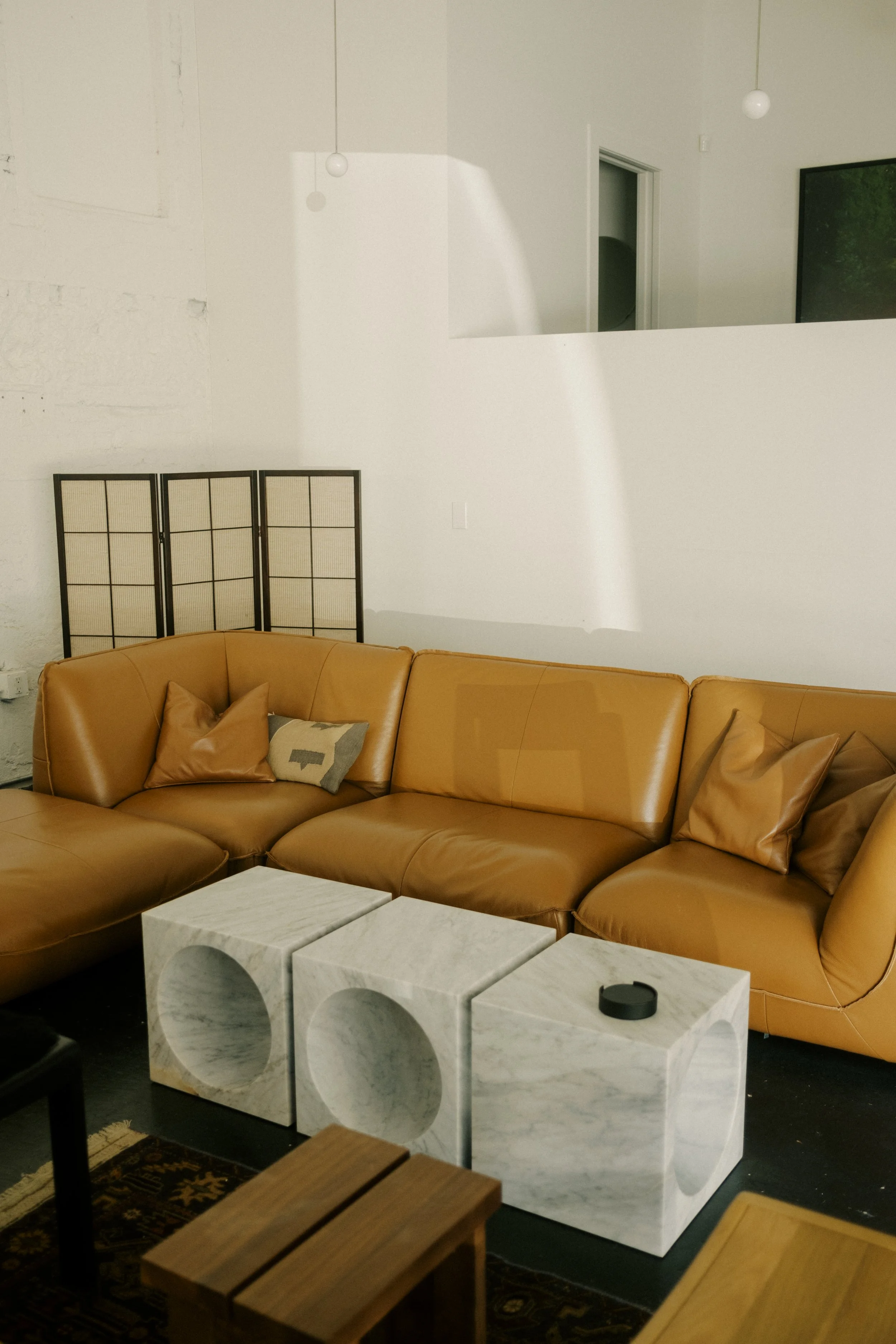 Living room with beige leather sectional sofa, pillows, marble cube tables, a black coffee table, a decorative folding screen, and a television on the wall.