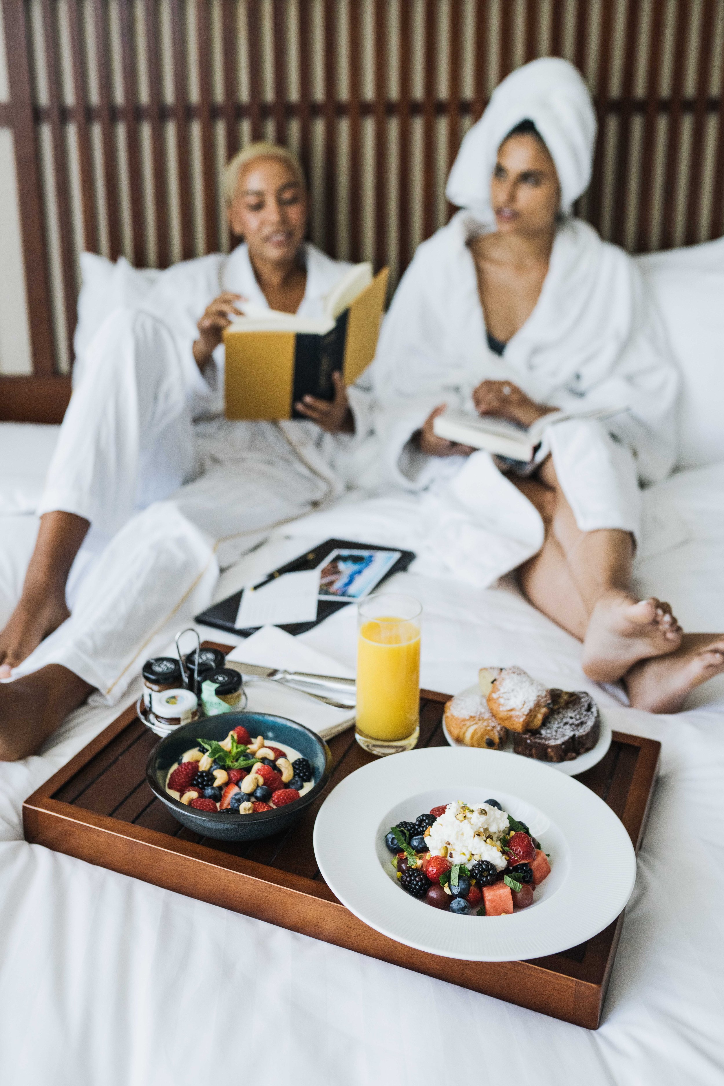 Two women in bathrobes sitting on a bed, reading books, with a breakfast tray in front of them that includes fruit, pastries, and orange juice.