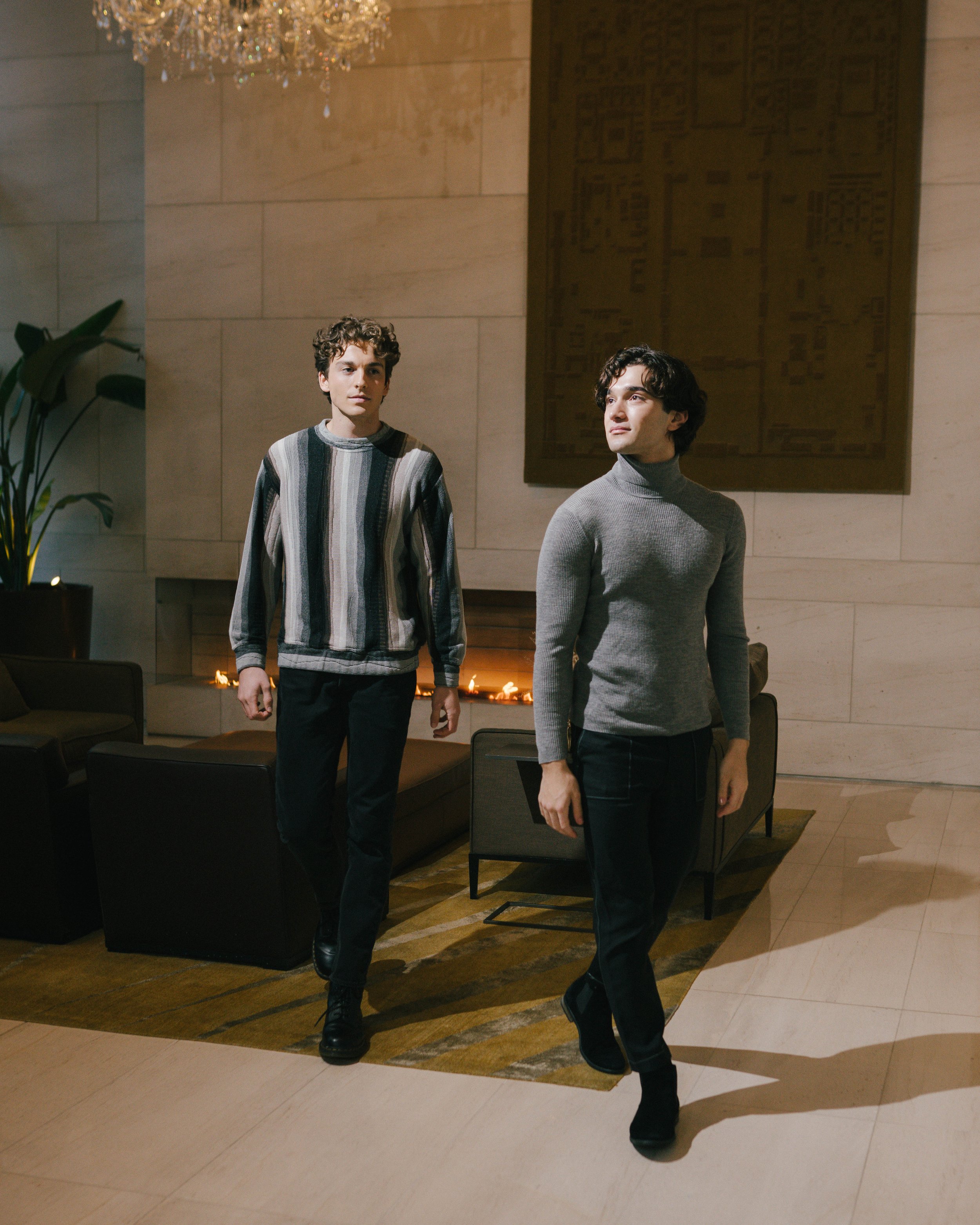 Two young men walking through a modern lobby with a fireplace, a large wall hanging, and elegant furniture.