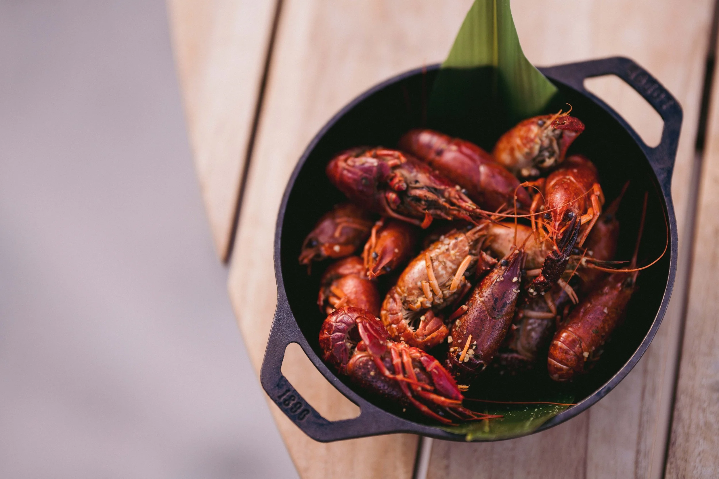 A black cast iron skillet filled with cooked crawfish placed on a wooden surface with a green leaf lining inside the pan.