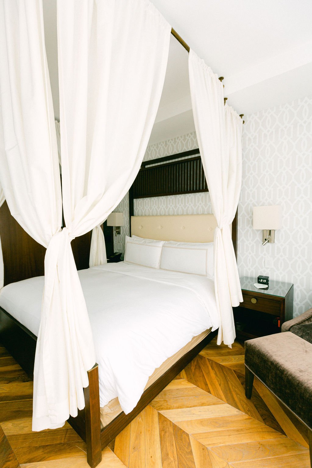 A hotel room with a four-poster bed with white curtains, a patterned wallpaper, a nightstand with a clock, a wall-mounted lamp, and a chair with brown upholstery.