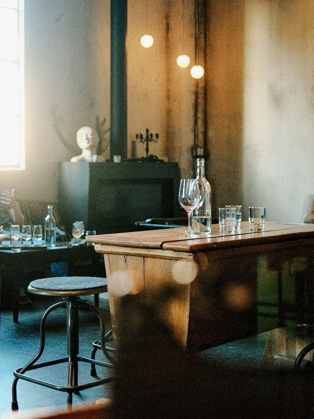 A rustic indoor setting with a wooden table, a glass bottle, wine glass, and shot glasses, with a vintage black radio, bust sculpture, and pendant lights in the background.