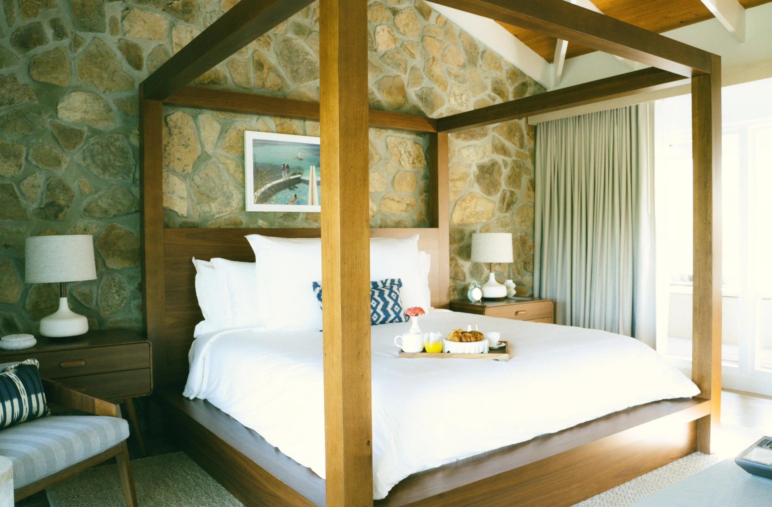 A cozy bedroom with a four-poster wooden bed, white bedding, and stone wall behind the headboard. There are two bedside tables with lamps, a framed picture on the wall, and a tray with breakfast on the bed.