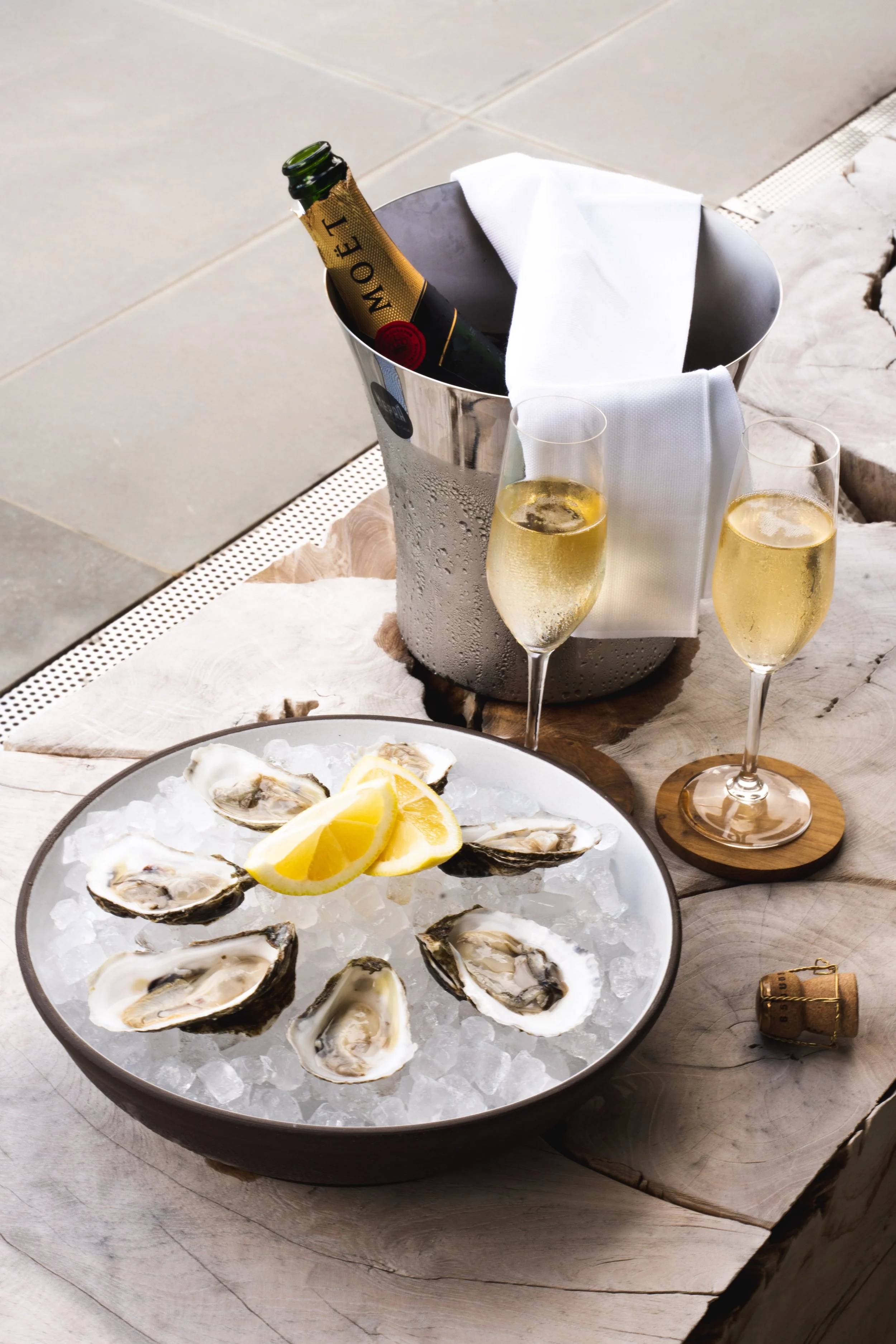 A bowl of fresh oysters on ice garnished with lemon wedges, two glasses of champagne, a bottle of champagne in an ice bucket with a white cloth, and a cork on a rustic wooden table.