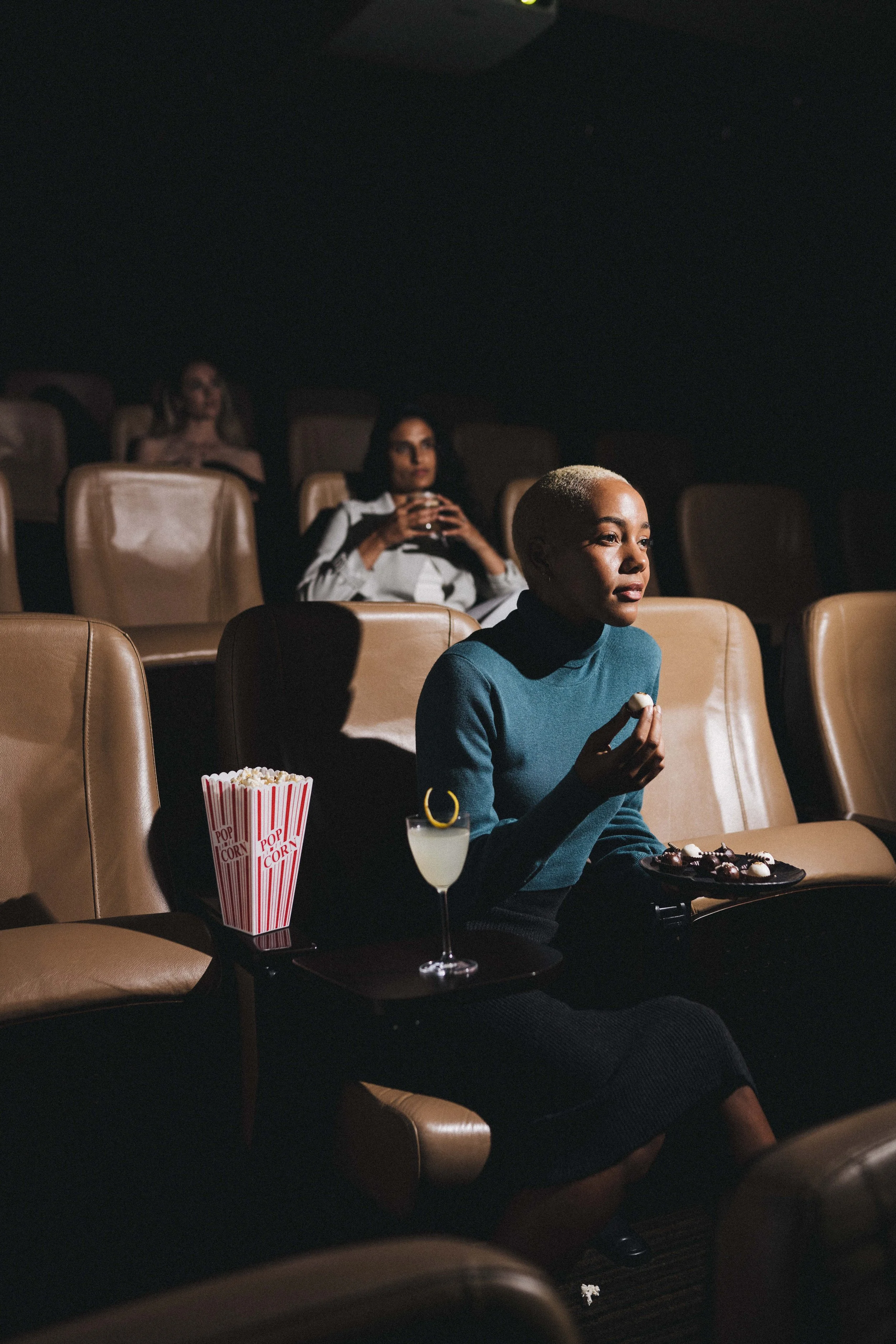 A woman with short blonde hair and a blue turtleneck is sitting in a movie theater, holding a piece of popcorn and a tray with chocolates, with a cup of popcorn and a drink on a small table beside her.