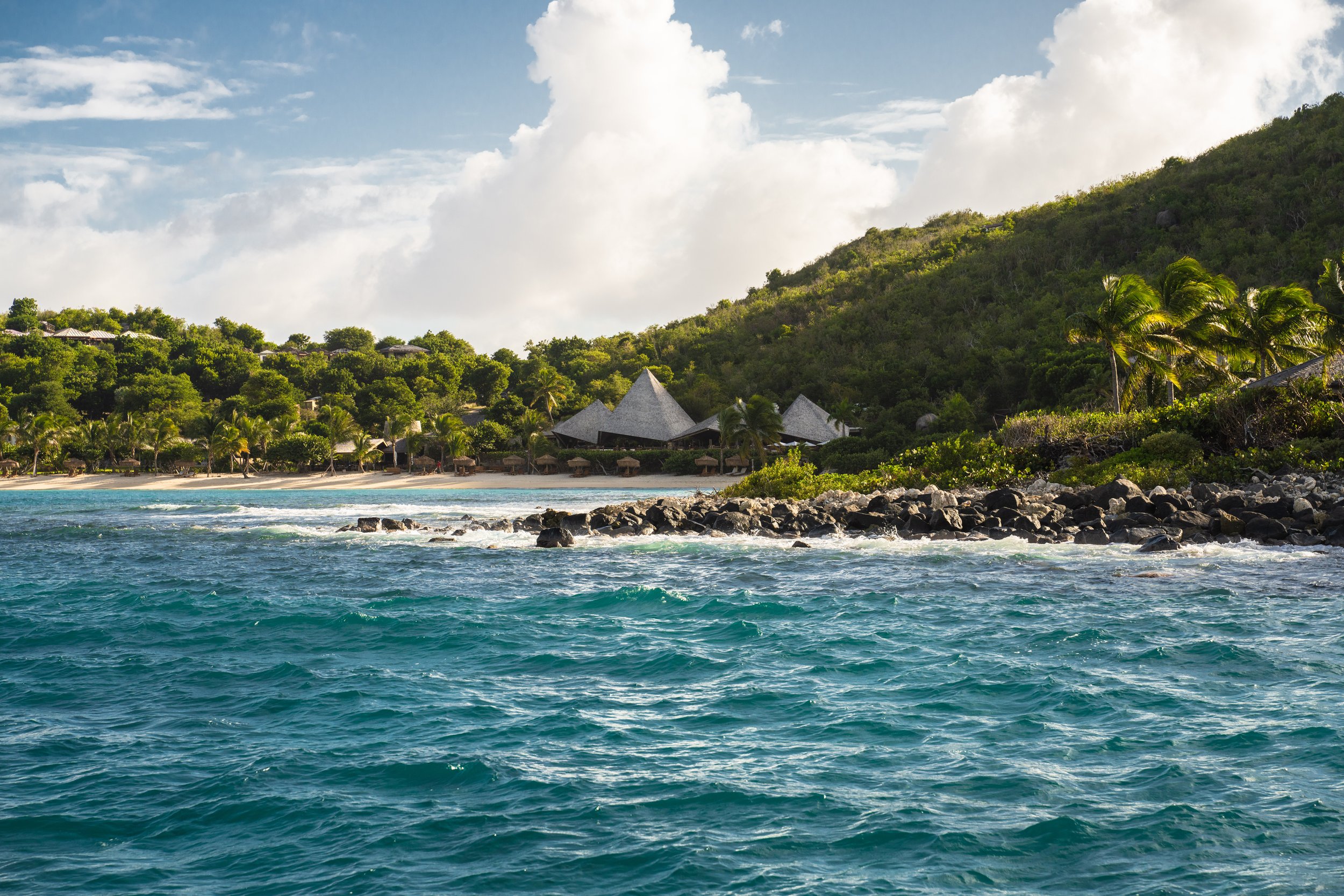 Tropical beach with turquoise water, rocky shoreline, palm trees, lush green hillside, and structures with thatched roofs under a partly cloudy sky.