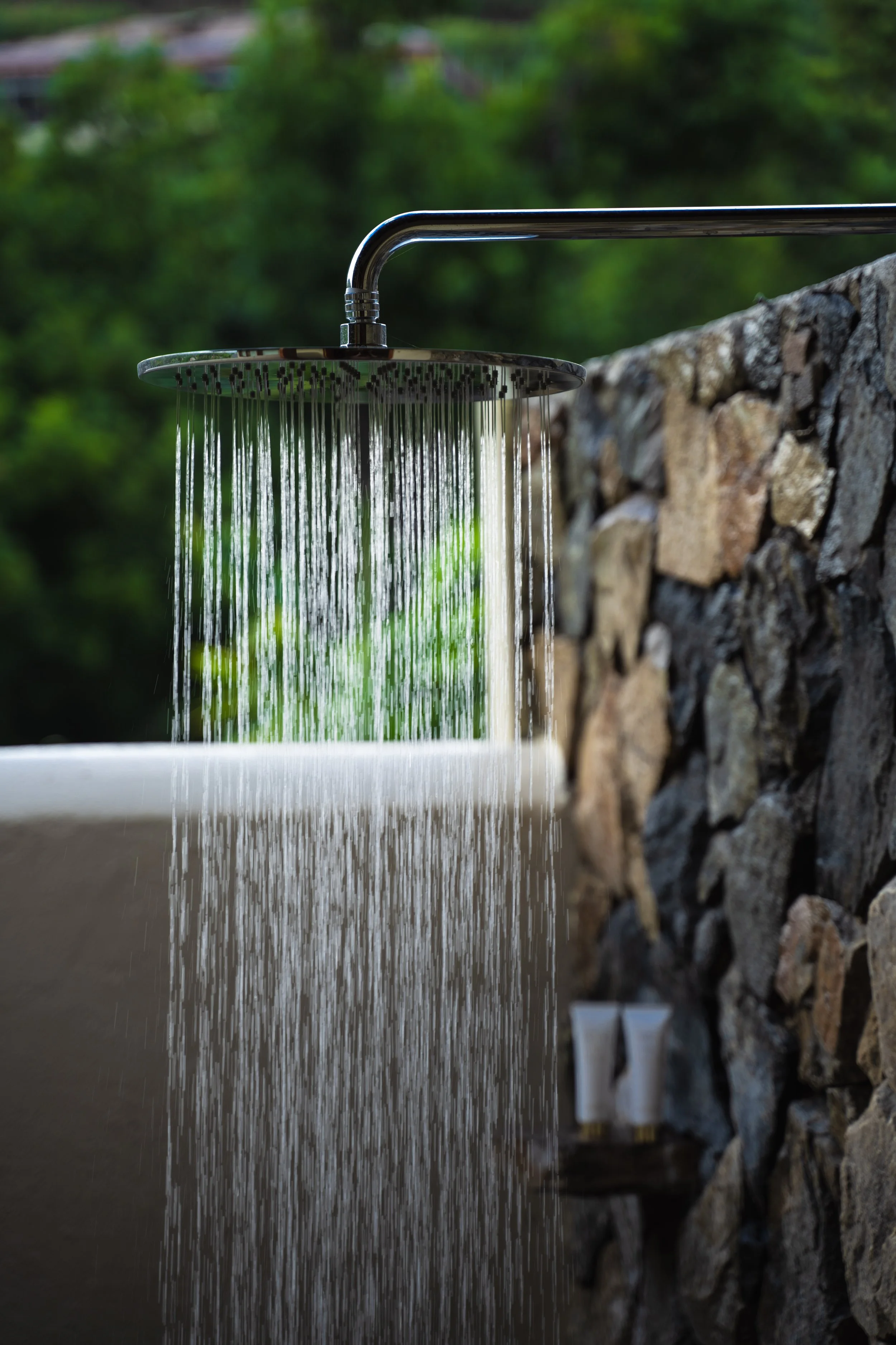 Outdoor shower with water flowing, mounted on a stone wall with greenery in the background.