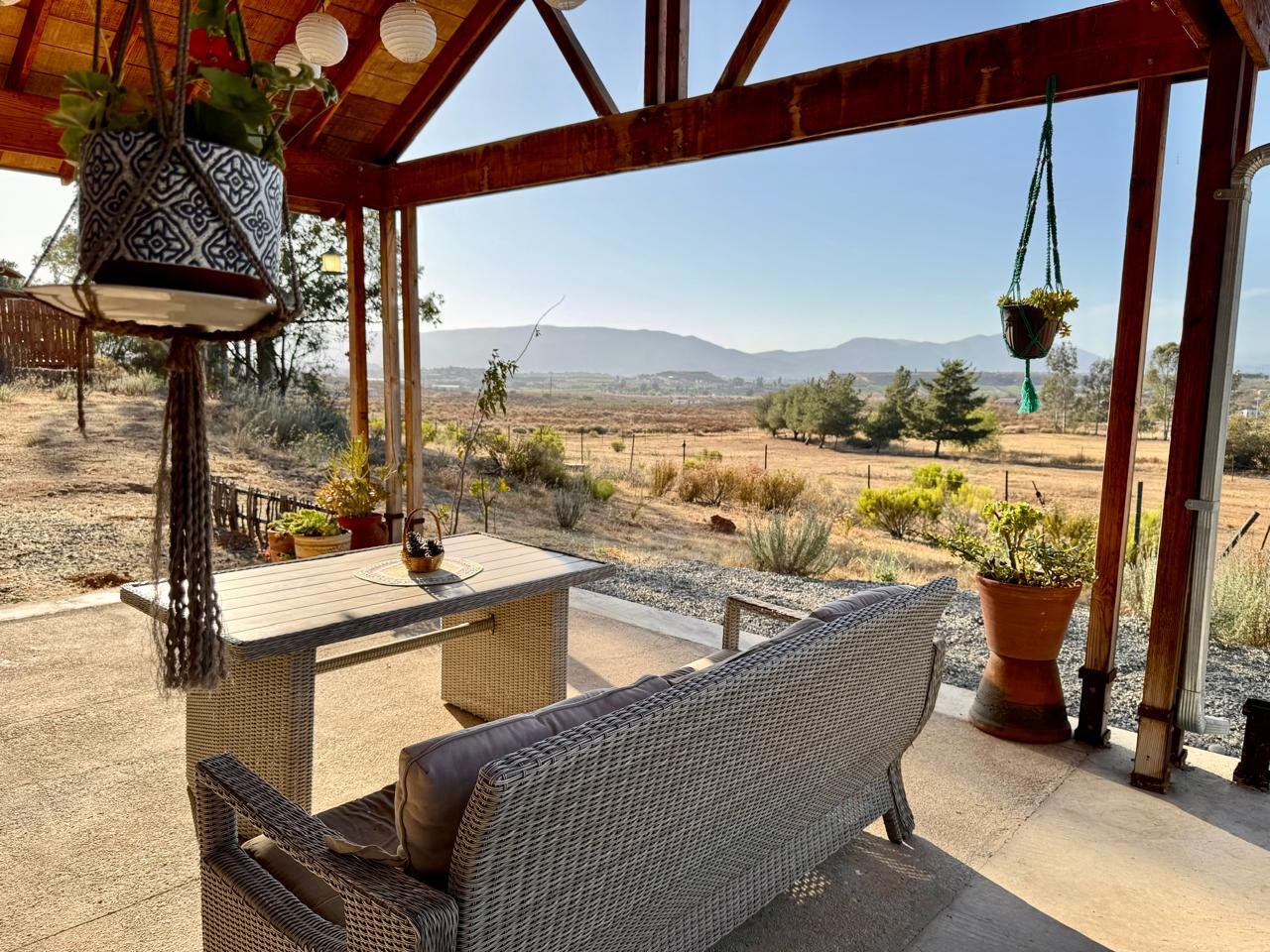 Vista de un porche con muebles de mimbre, mesa y plantas en macetas, que mira hacia un campo árido y montañas al fondo, en un día soleado.