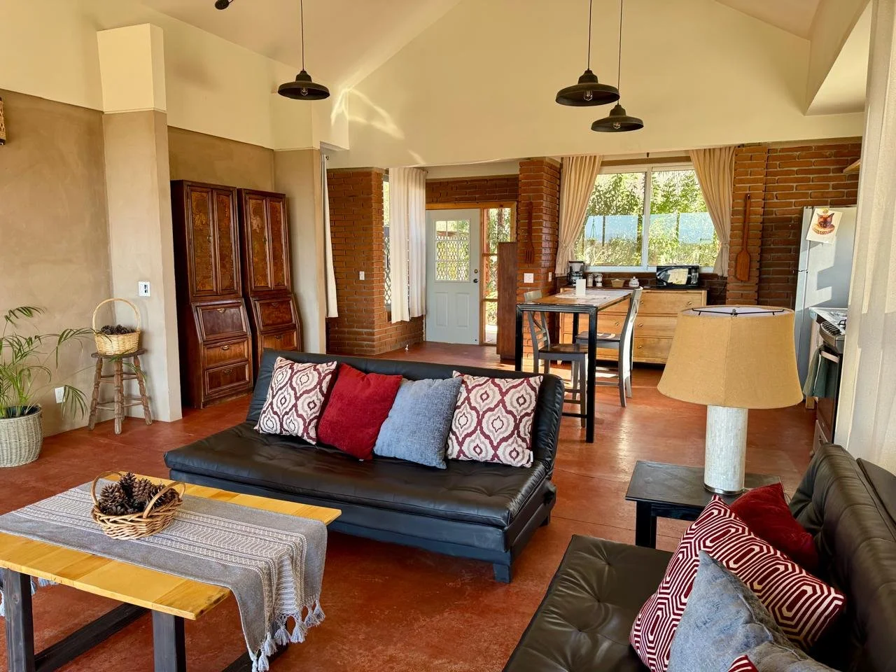 Sala de estar con muebles de piel negra, cojines decorativos, lámpara de mesa, mesa de café con cesta y piñas, cocina en el fondo con ventanas enmarcadas por cortinas beige, paredes de ladrillo expuesto y piso de madera.