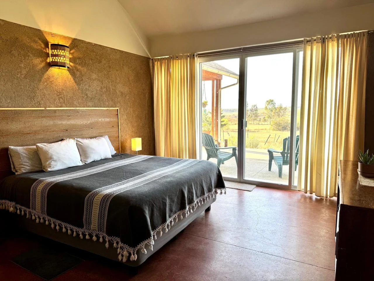 Habitación con cama grande, cortinas amarillas, puertas corredizas que dan a un balcón con dos sillas, vista hacia naturaleza con árboles y campo.