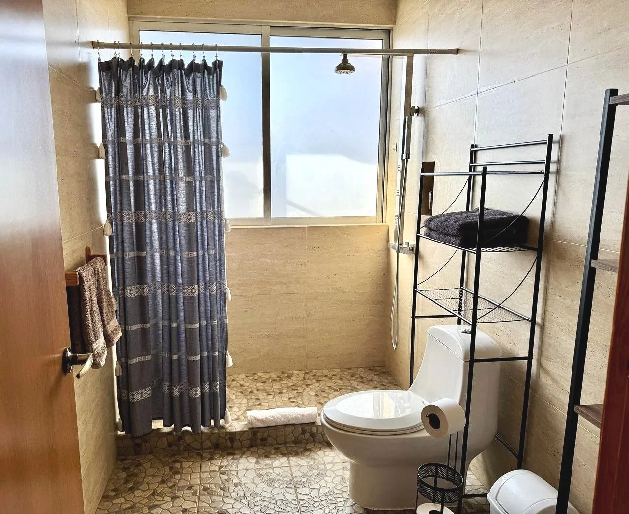 Baño con pared de azulejo beige, ducha con cortina azul, escalera metálica con toallas y ropa, inodoro con papel higiénico, ventana grande con cortina, suelo de piedra, escoba y cubo de basura.