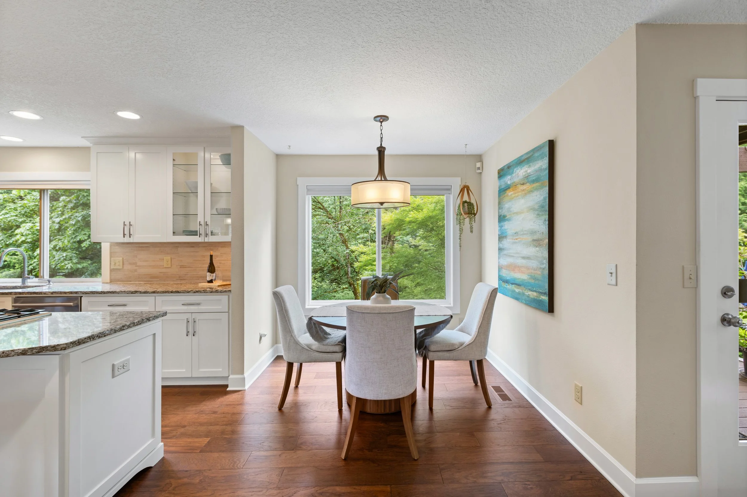 Dining area with a round table, four upholstered chairs, large window with greenery outside, artwork on wall, and a hanging light fixture.