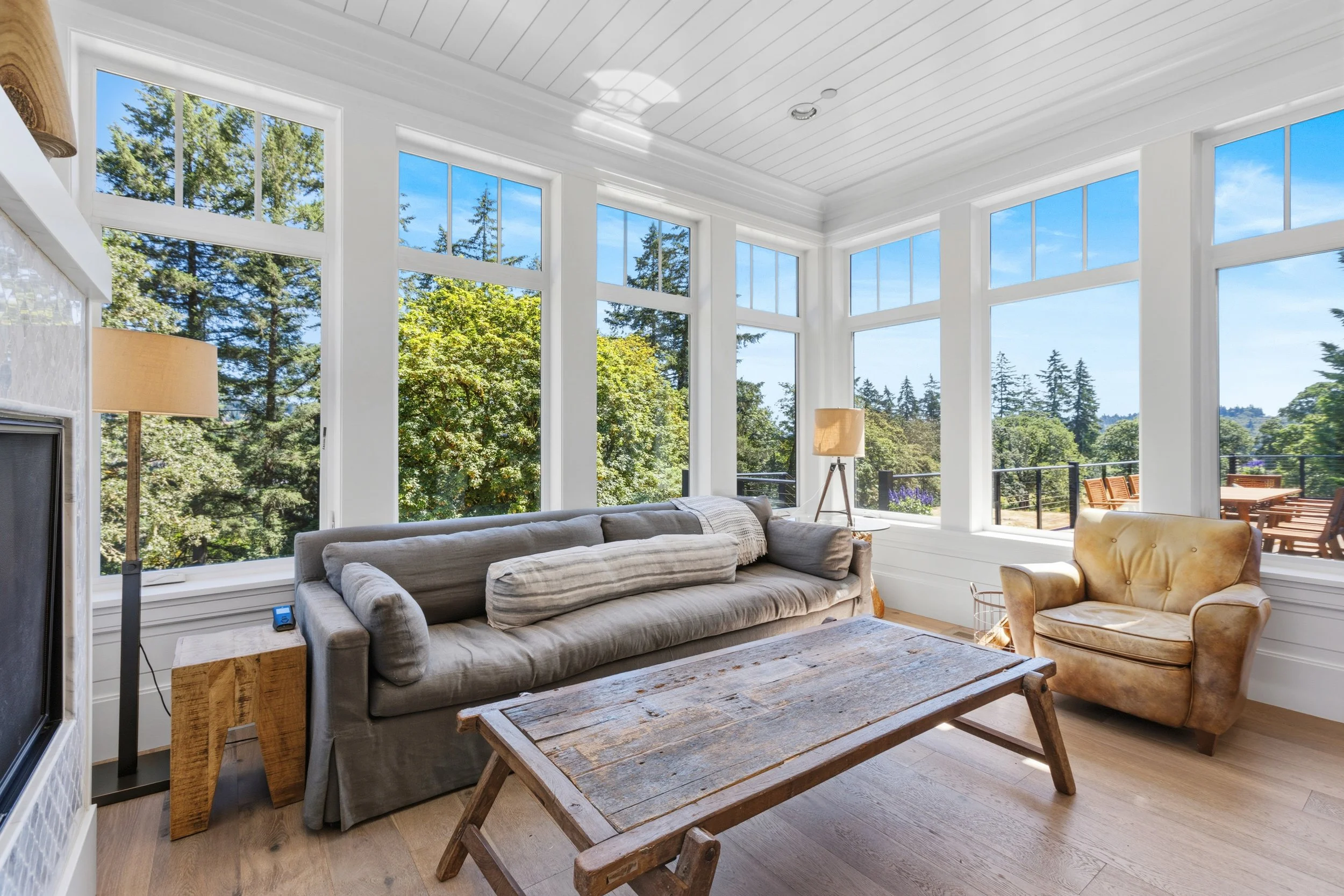 A living room with large windows overlooking trees and a deck, featuring a grey sofa, a wooden coffee table, a beige armchair, and a floor lamp with a beige shade.