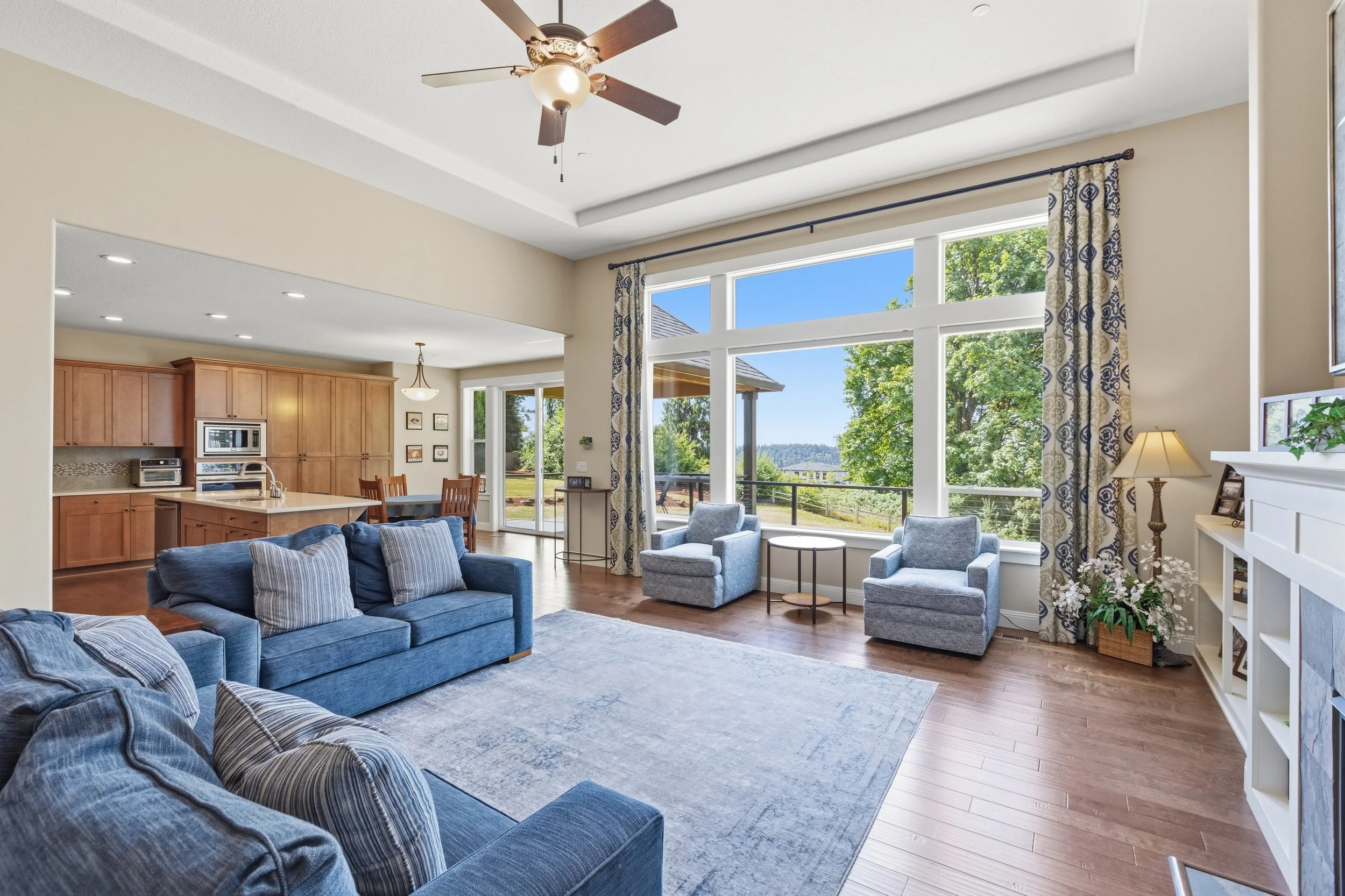 Living room with large windows, blue sofa, armchairs, hardwood floors, and view of green trees outside.