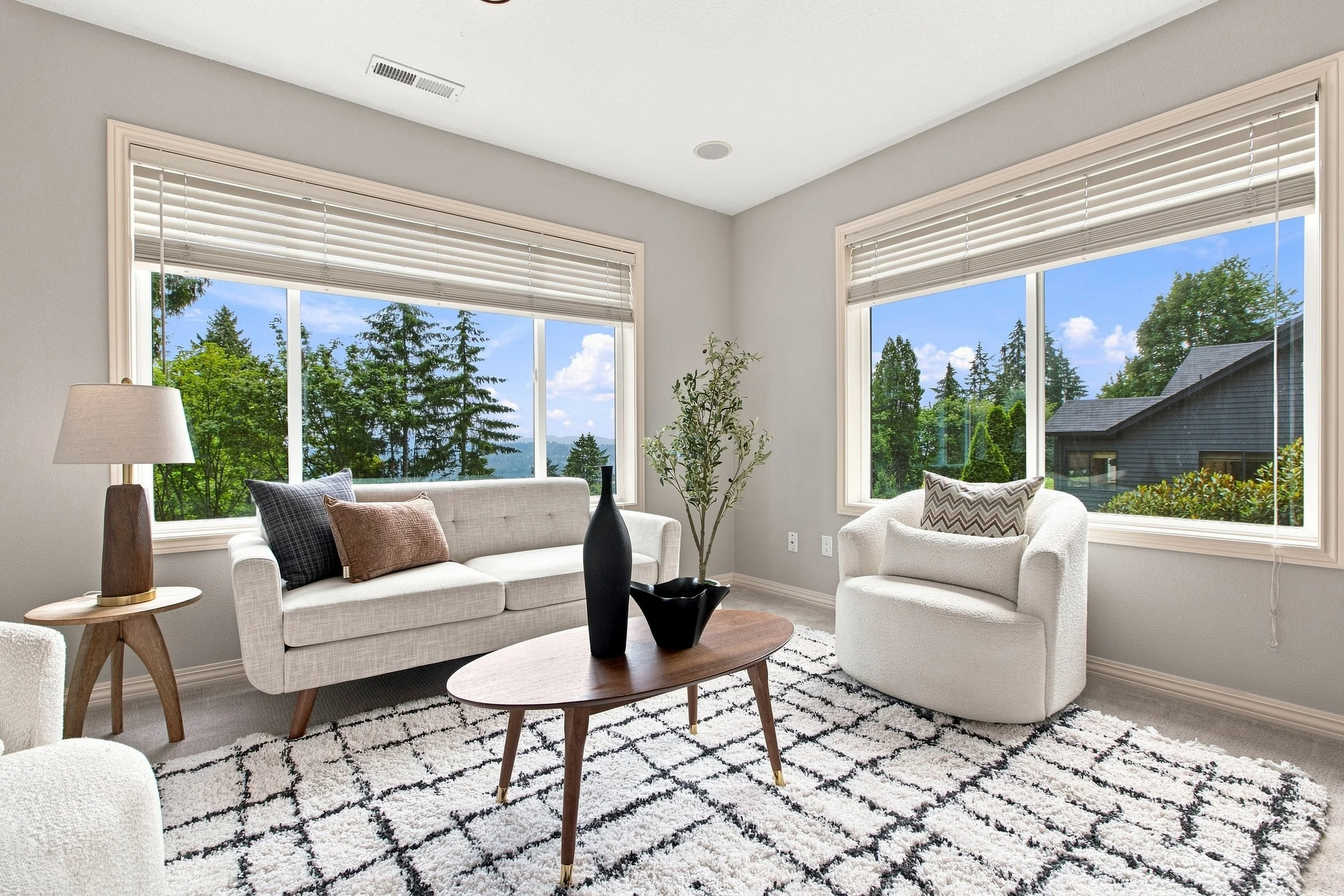 Living room with large windows showing a view of trees and blue sky, furnished with a white sofa, armchair, wooden coffee table, throw pillows, potted plant, lamp, and a black vase, with a black-and-white patterned rug.