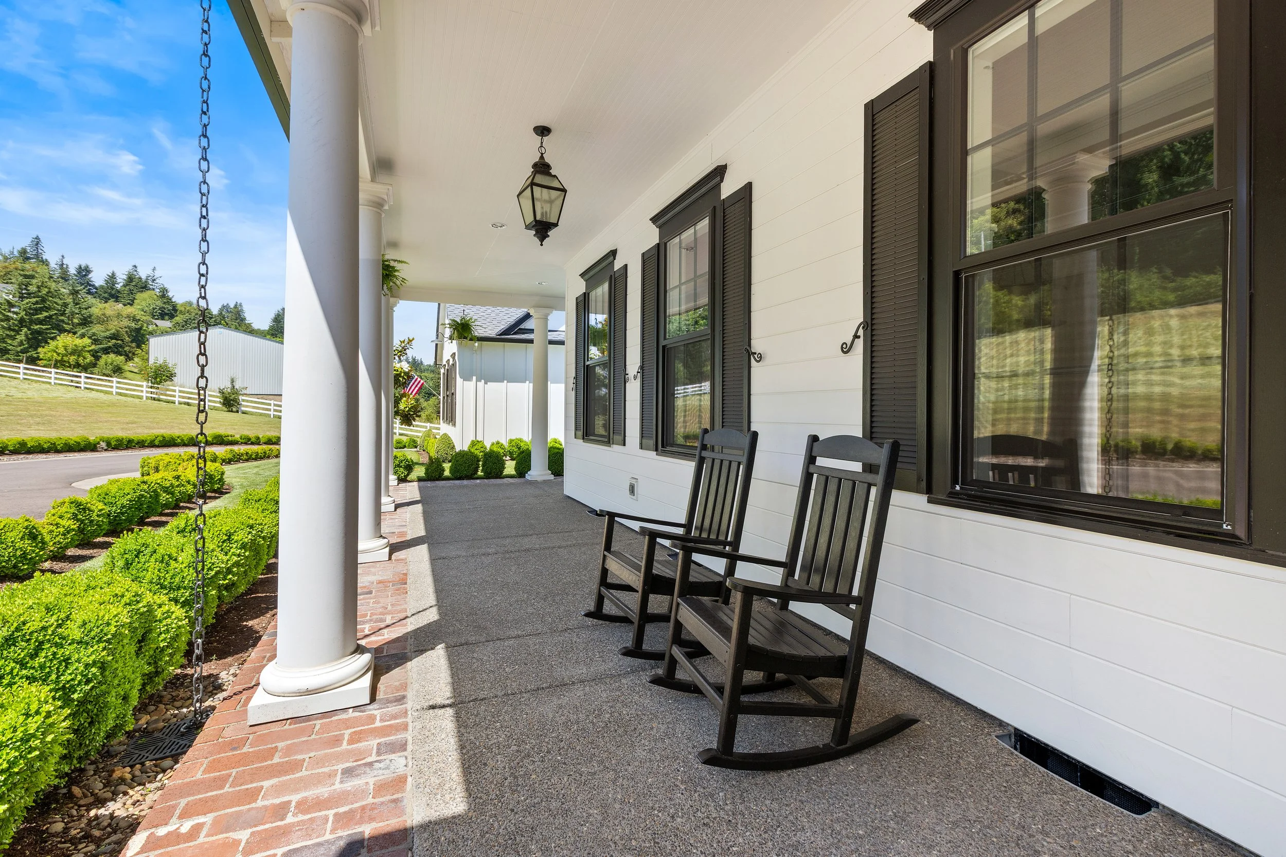 Front porch of a house with black window shutters, white siding, a hanging lantern light, and wooden chairs. There are columns supporting the porch roof, a brick walkway, and neatly trimmed bushes in the yard. The sky is clear and blue, and the backg