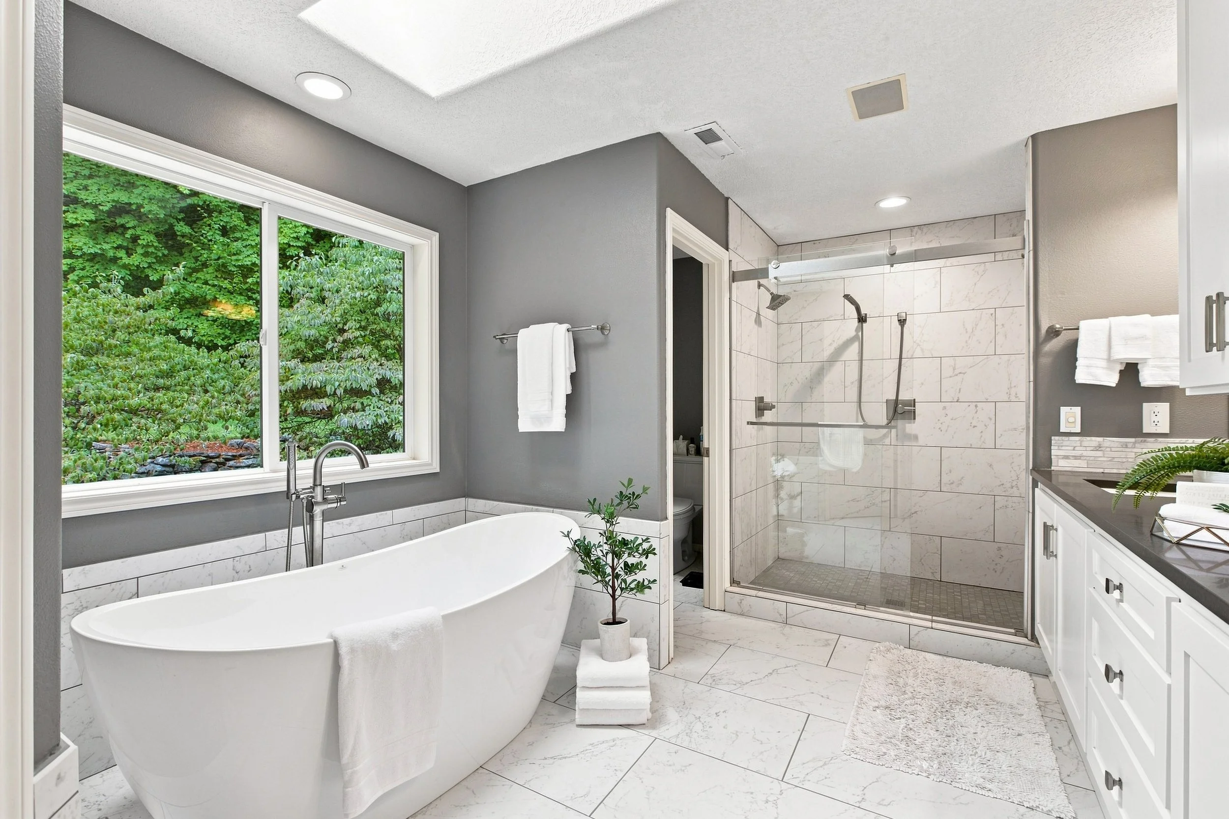 Modern bathroom with large window showing green trees outside, freestanding bathtub, walk-in shower with marble tiles, bathroom vanity with sink, white towels, and a small potted plant.
