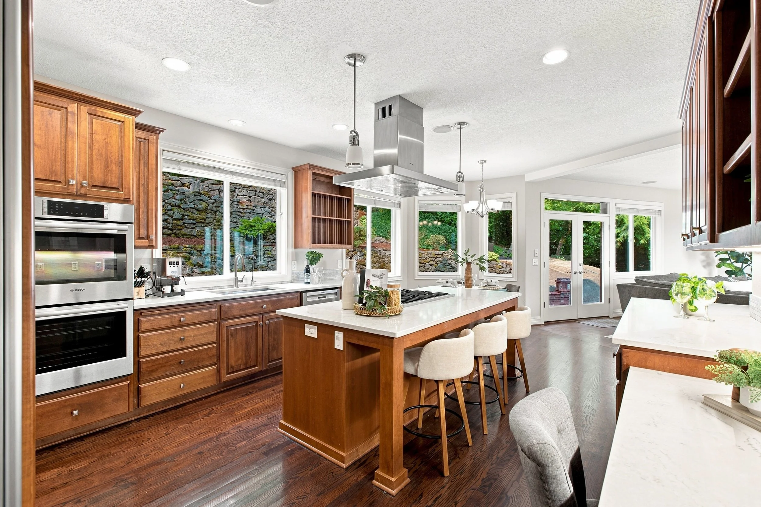 Kitchen with wooden cabinets, white countertops, and a central island with bar stools. Large windows overlook a stone wall and greenery outside, allowing natural light into the space. There's a stainless steel range hood hanging over the island and d