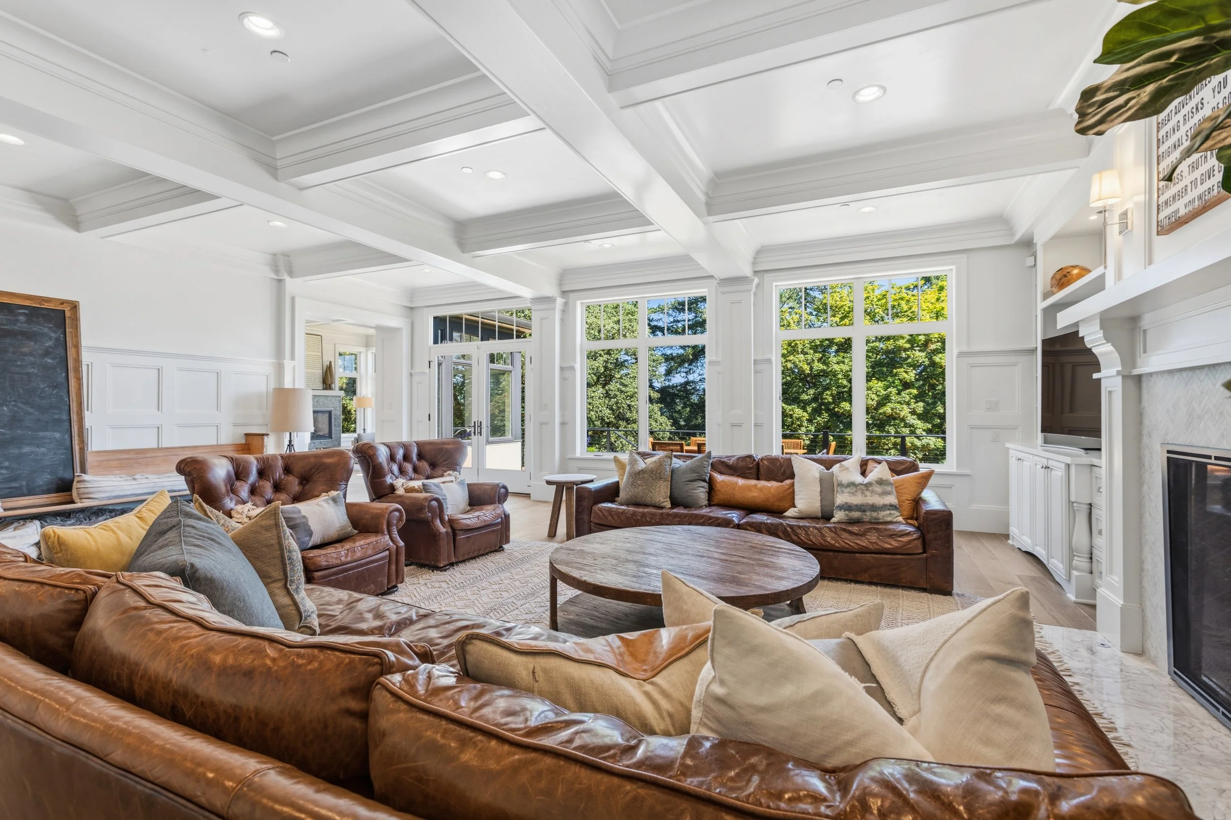 Living room with large windows, white walls, and a white coffered ceiling. Features brown leather sofas with various pillows, a round wooden coffee table, and a fireplace with built-in shelves. There are trees visible outside through the windows.
