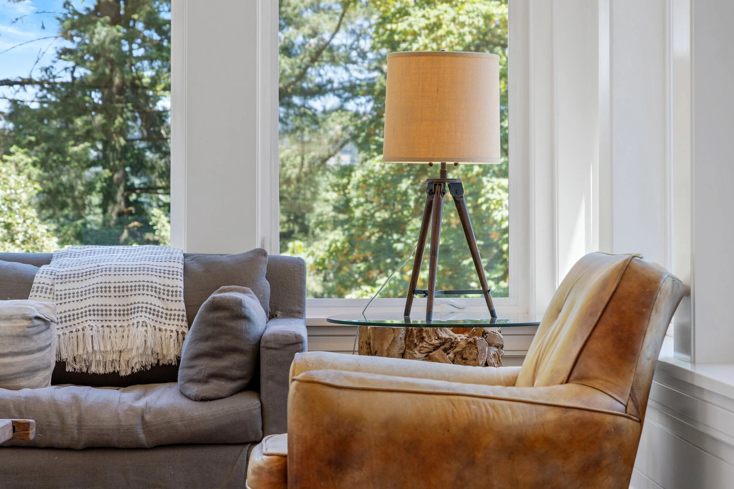 A cozy living room with a beige sofa covered with a textured throw, a wooden side table made from a broken tree trunk, a tripod-style floor lamp with a beige lampshade, and a large window showing green trees outside.