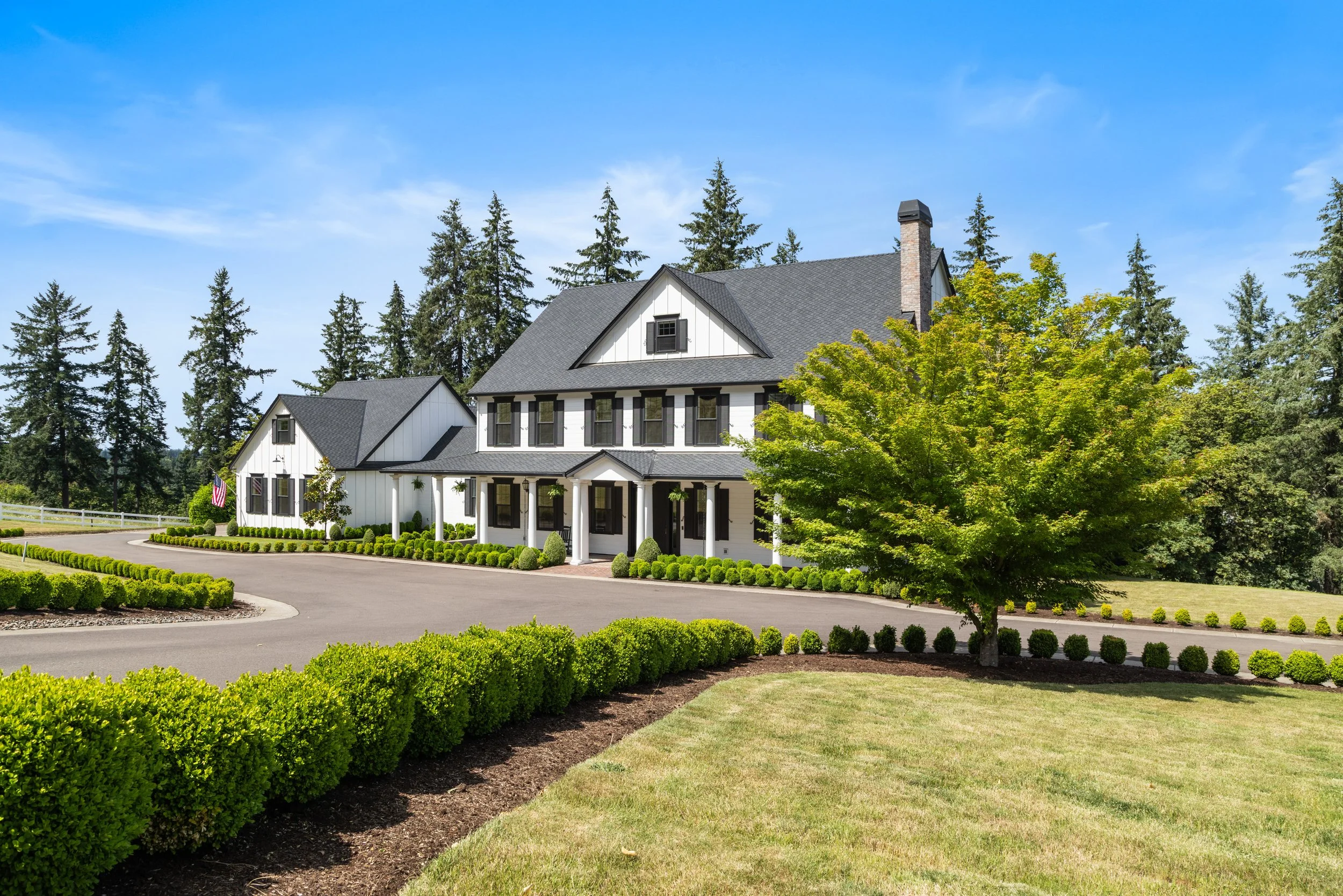 A large white house with black shutters and a dark roof, surrounded by green trees and well-maintained landscaping, under a bright blue sky.
