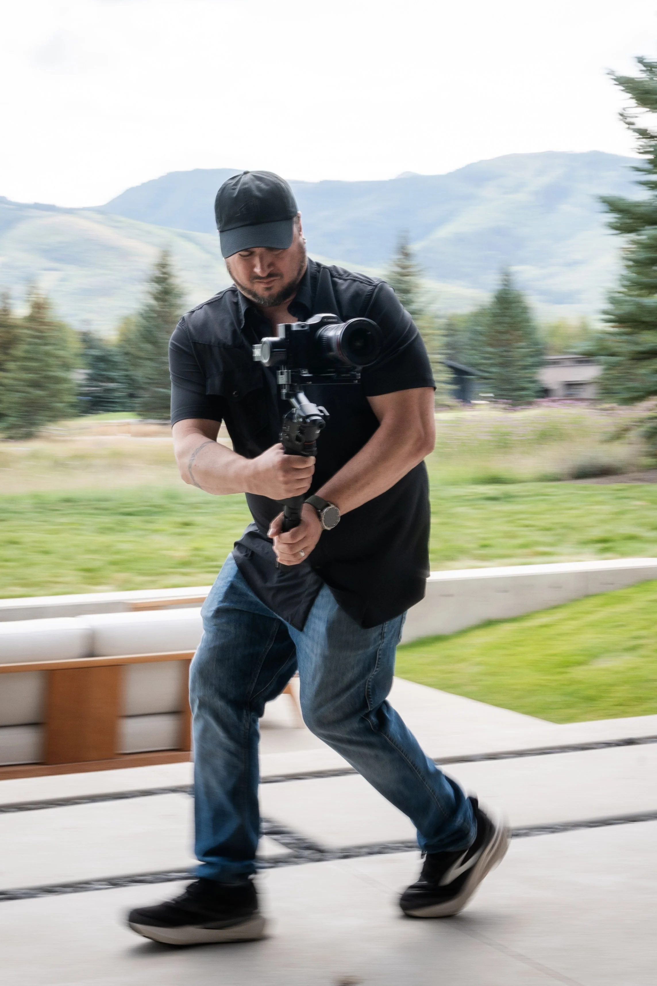 A man wearing a black cap and black shirt is holding a camera stabilizer while walking outdoors with mountains and trees in the background. John Cherniack, founder of Northwest Realty Media in action shoot video with his gimbal.