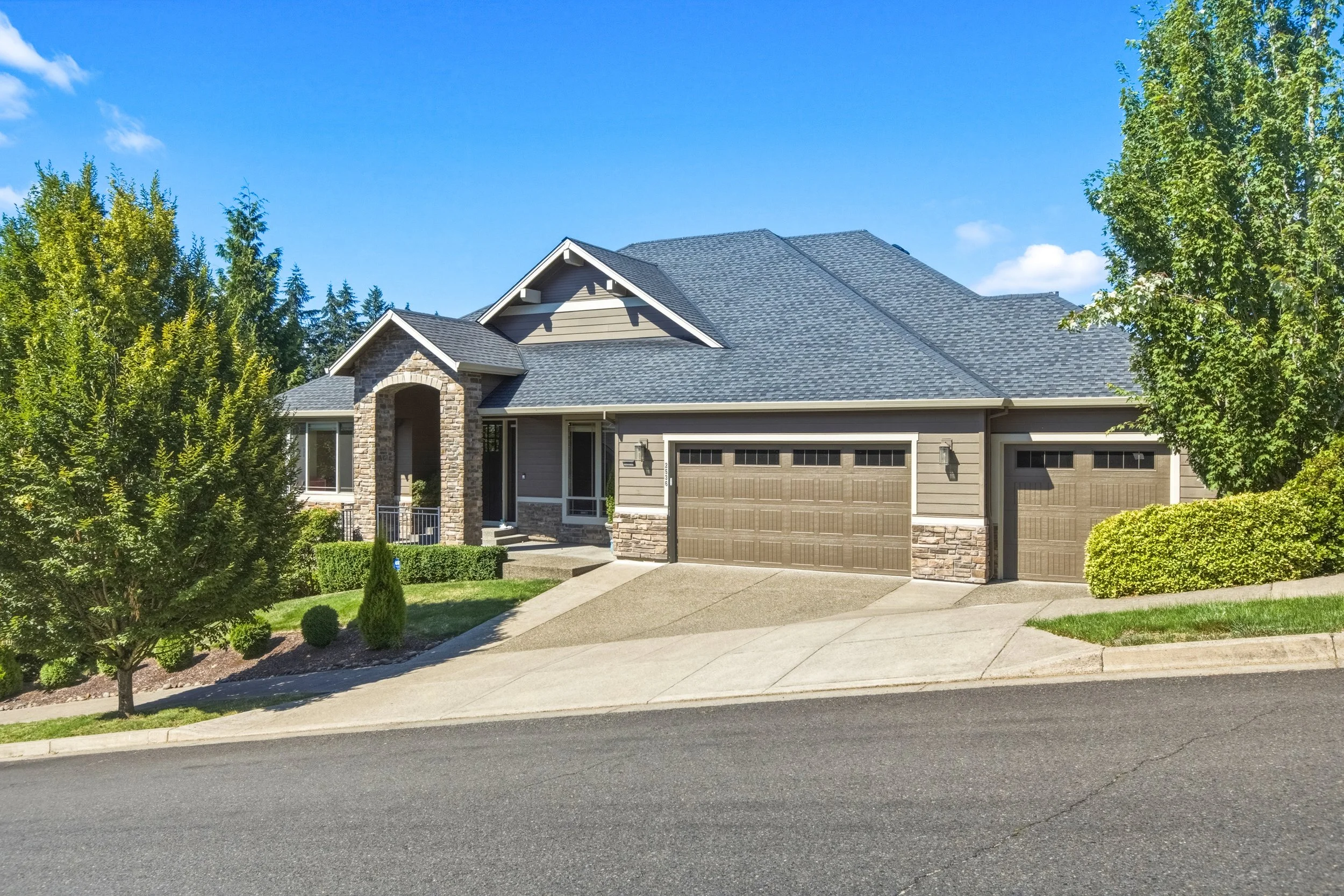 A modern house with a gray roof, stone and siding exterior, and a two-car garage. The house is surrounded by green trees and shrubs, with a paved driveway and a sidewalk in front. The sky is mostly clear with a few clouds.