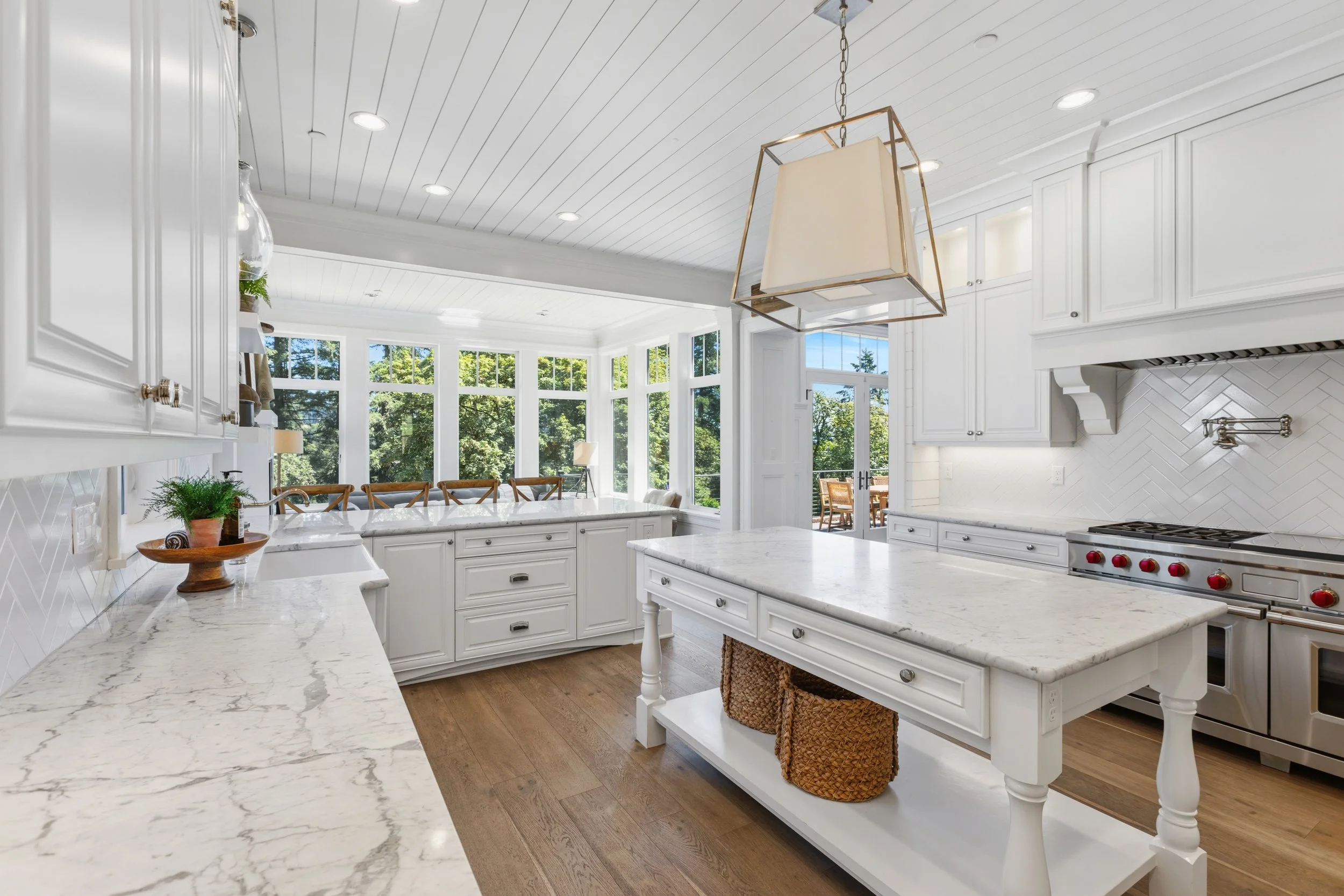 Bright, white kitchen with marble countertops, white cabinets, and large windows showing trees outside. A hanging light fixture and a door lead to an outdoor patio.