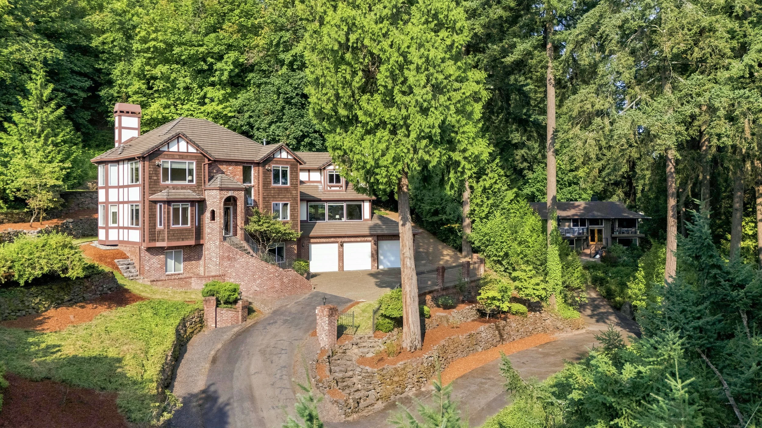 A large, multi-story house with a brick and wood exterior, surrounded by tall green trees. The house has a driveway leading to a garage and a landscaped yard.