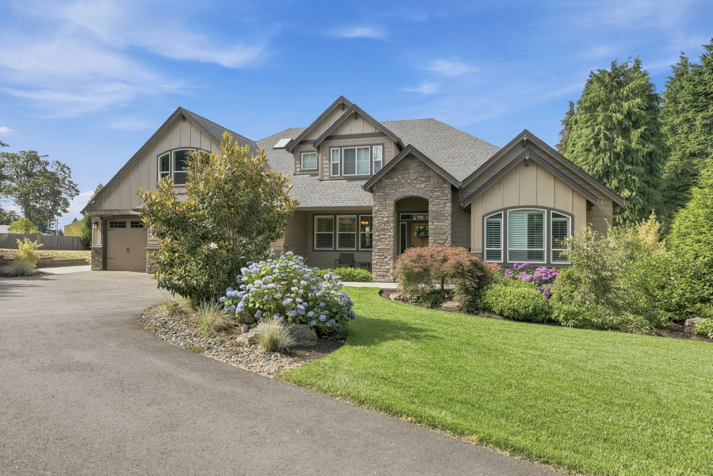 A two-story house with a landscaped front yard featuring various flowering plants and trees, under a blue sky with a few clouds.