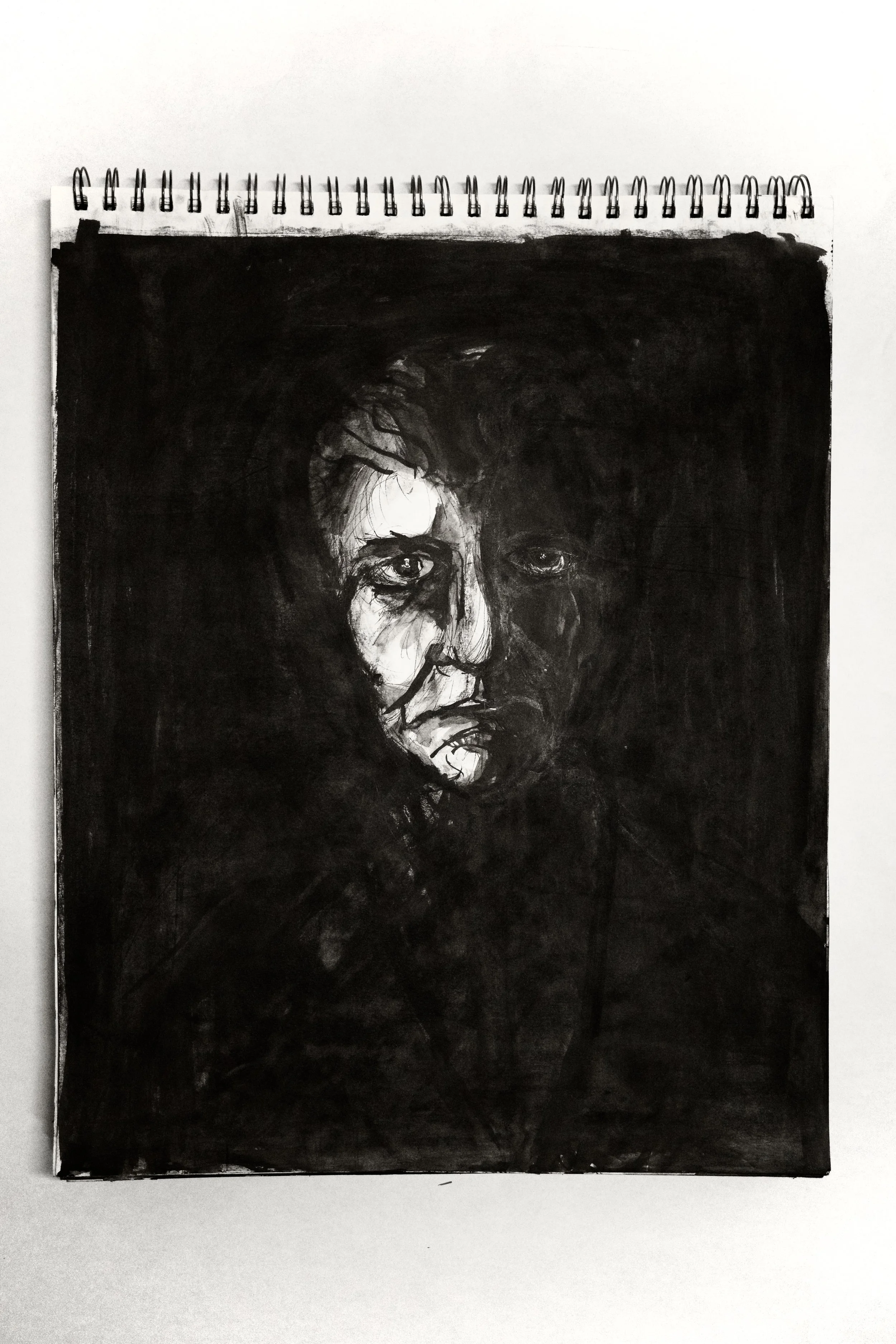 A black and white drawing of a person's face, with expressive eyes and dark shading around the features, on a spiral-bound sketch pad.