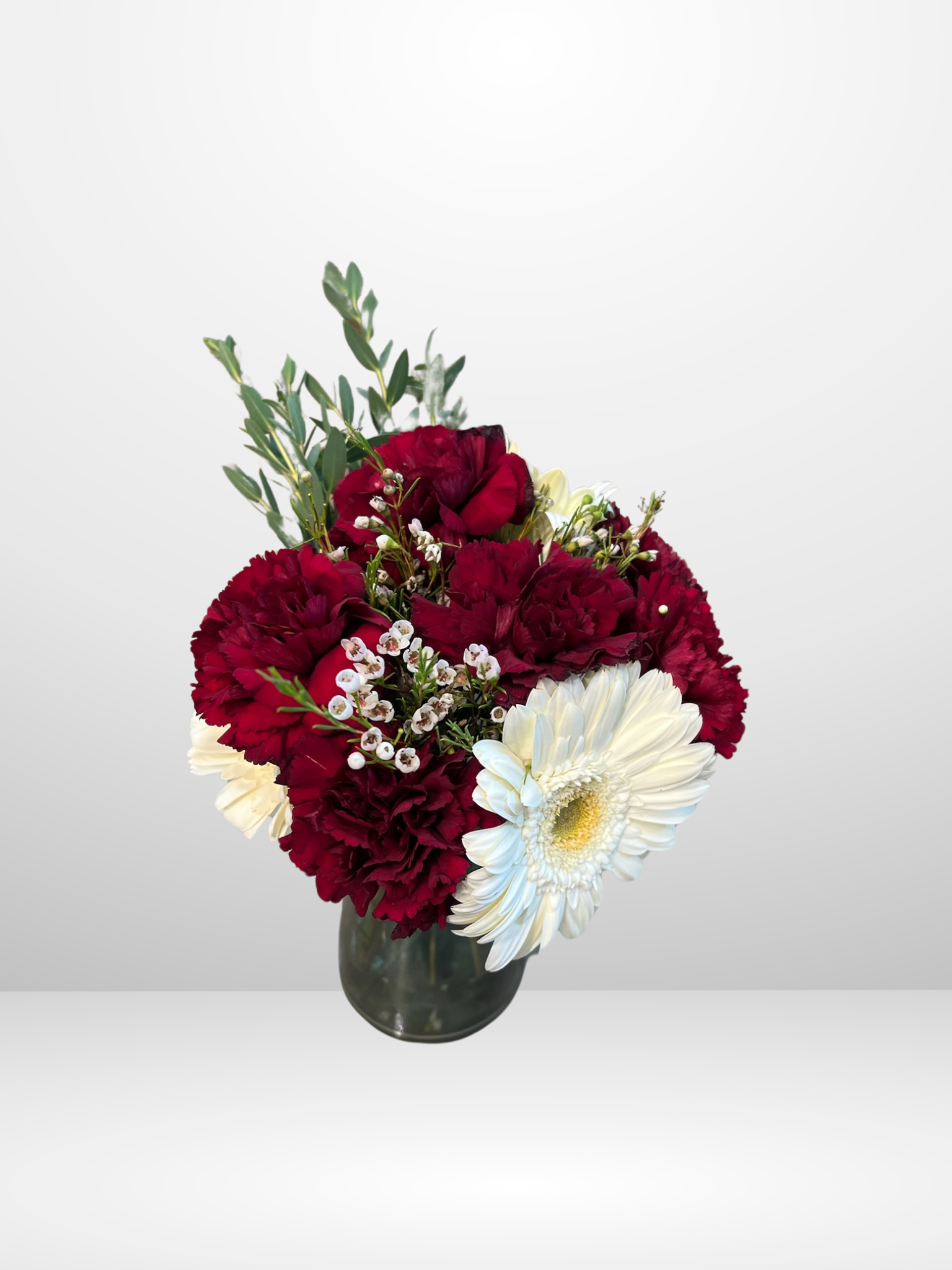A bouquet in a vase with red carnations, a white gerbera daisy, and green foliage on a light background.