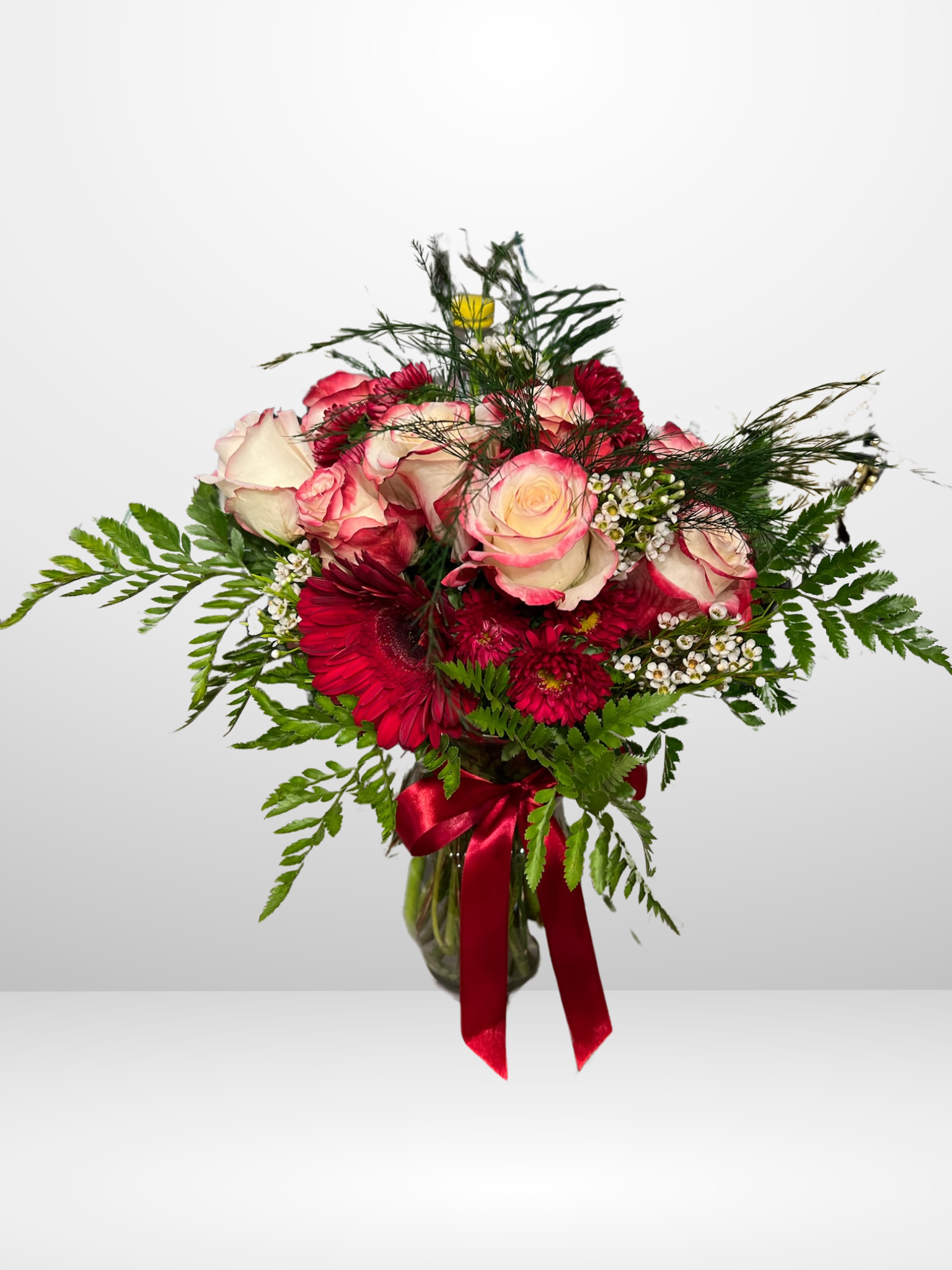 Flower bouquet with pink roses, red flowers, greenery, and a red ribbon in a vase.